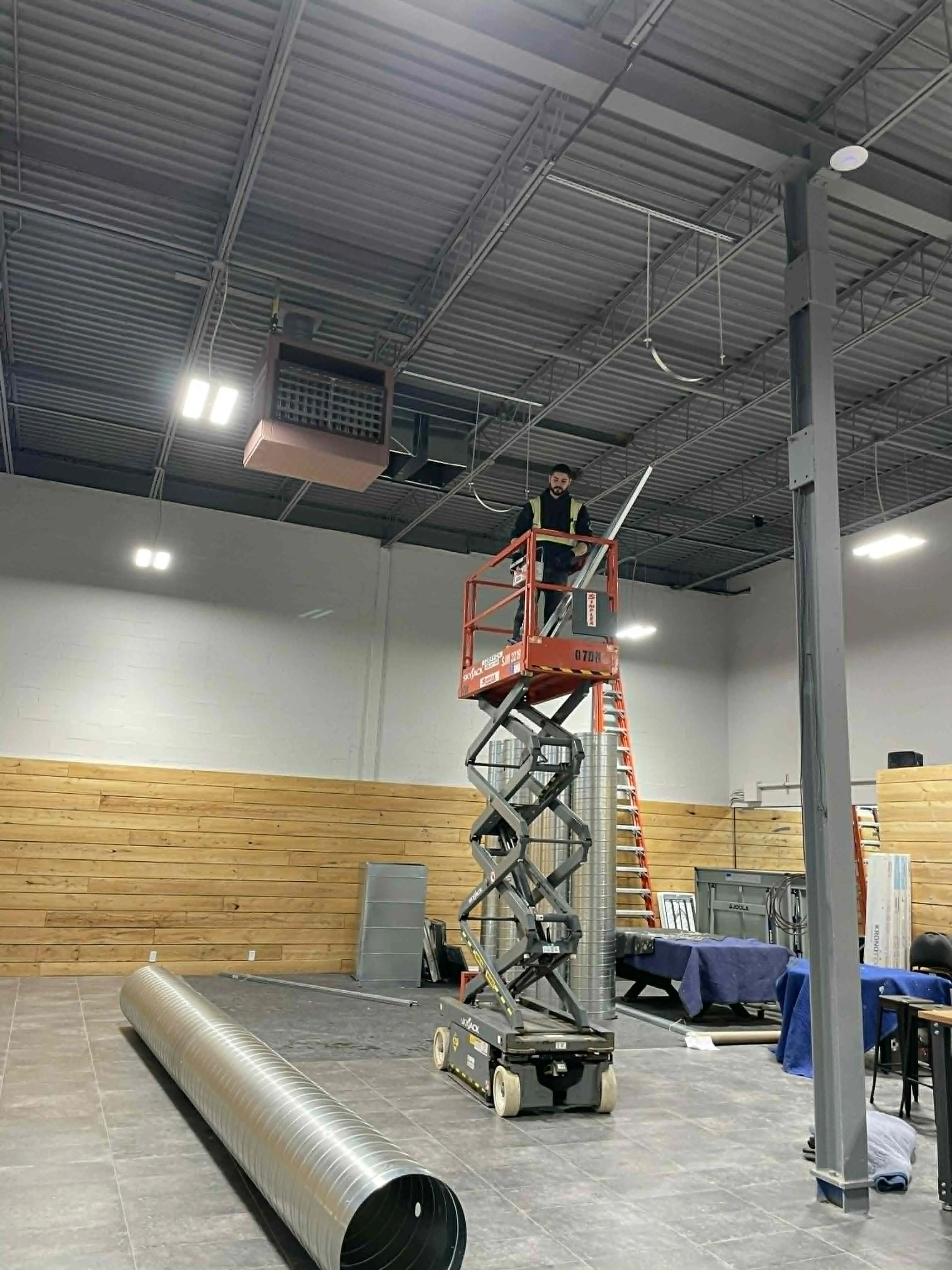 Une personne travaillant en hauteur dans un espace industriel, utilisant une plateforme élévatrice pour manipuler un tuyau en métal suspendu au plafond. La pièce a un plafond en métal apparent, avec un mur en bois et une zone de sol en gres.