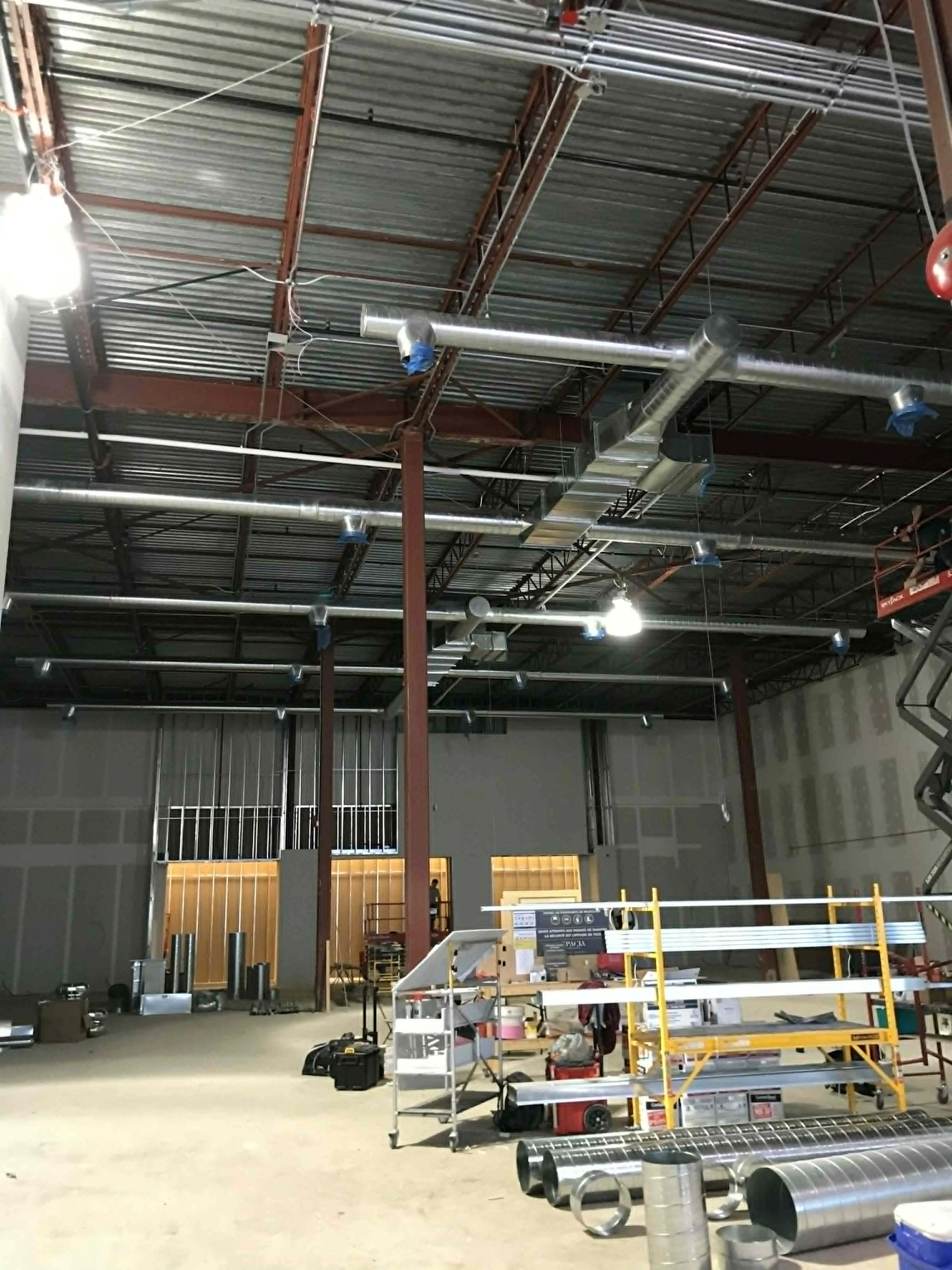 Intérieur d'une construction industrielle avec des conduits en métal suspendus au plafond, des outils, des équipements, un échafaudage et des matériaux de chantier au sol.
