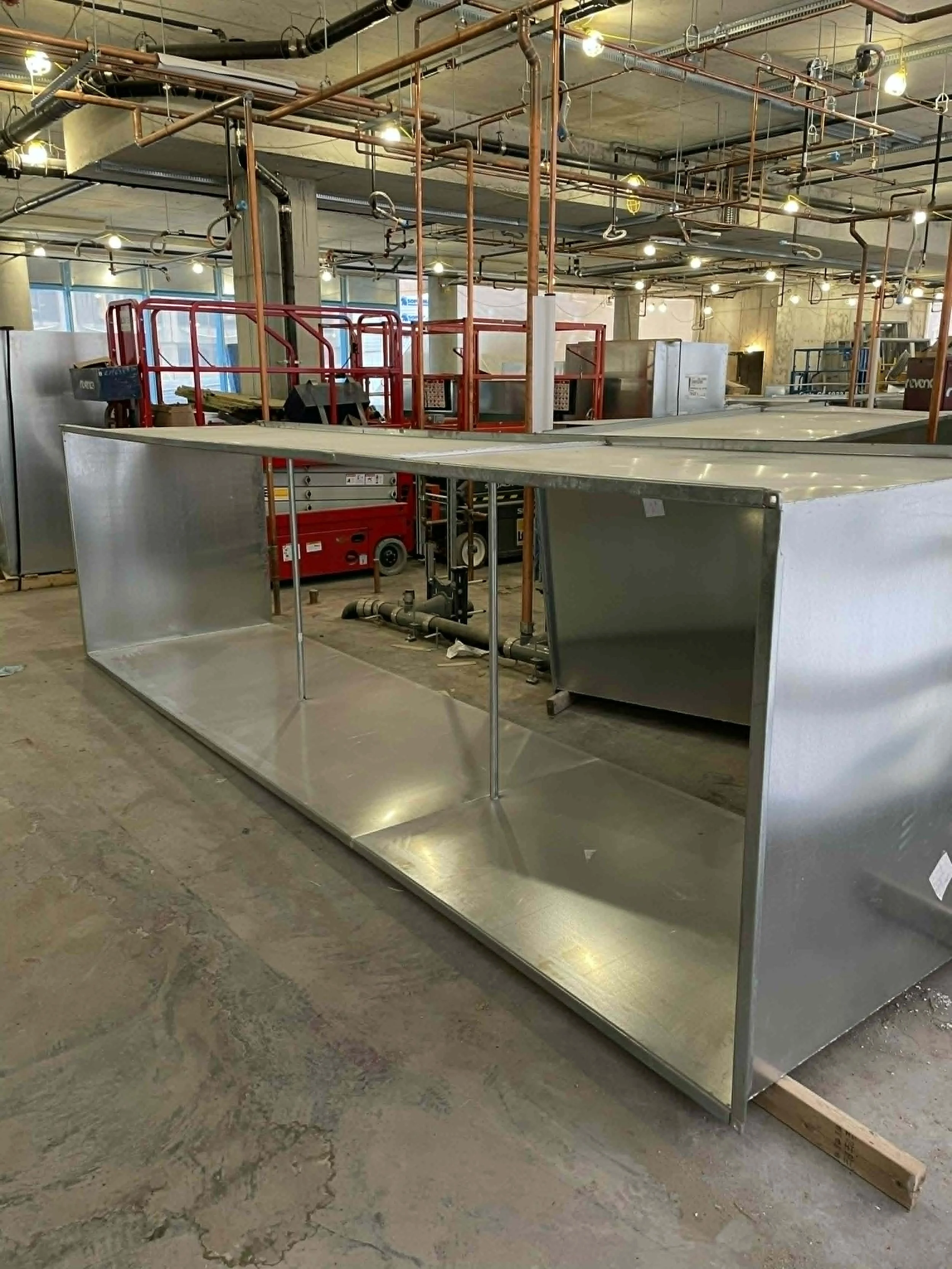 Une grande structure en acier inoxydable en cours d'installation dans un espace intérieur industriel, avec un réseau de tuyaux suspendus au plafond et des lumières encastrées.
