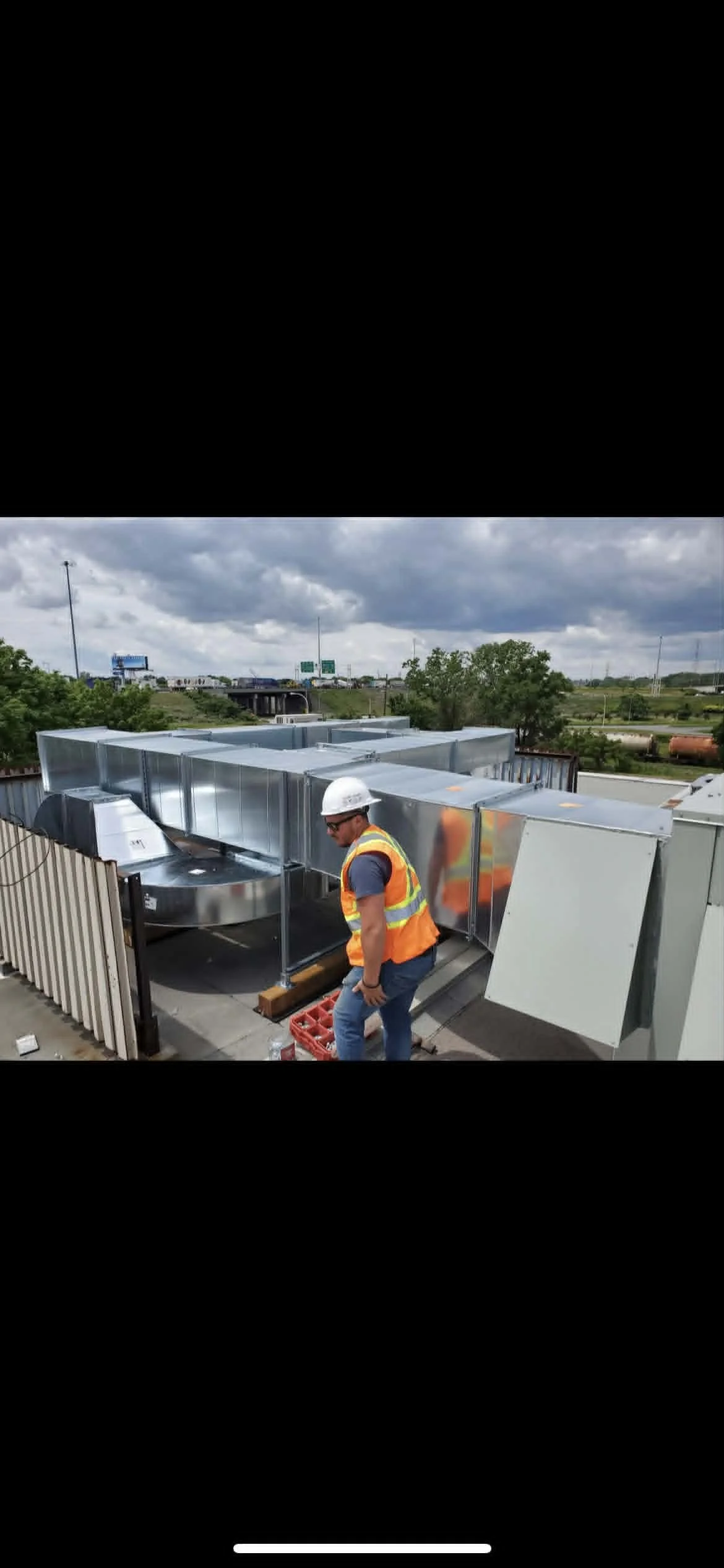 Un homme portant un gilet de sécurité orange et un casque blanc travaille sur un toit en utilisant des outils, avec des conduits de ventilation en métal autour de lui.