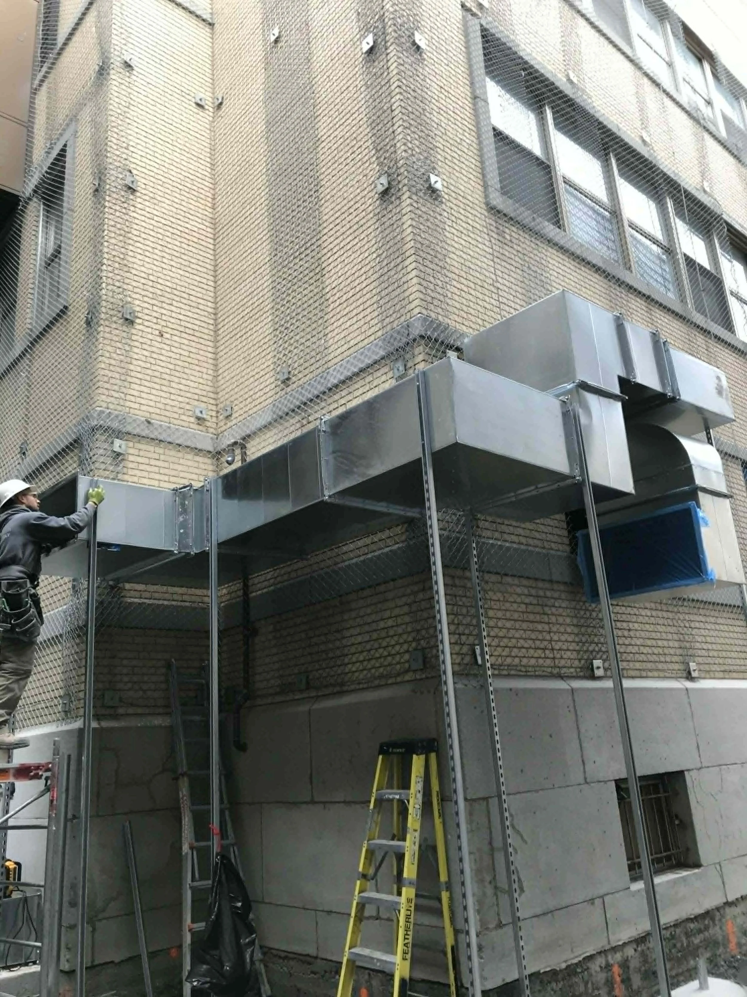 Travaux de rénovation ou d'installation sur la façade d'un immeuble en briques, avec un échafaudage métallique, un homme en casque et gants, un escabeau jaune.