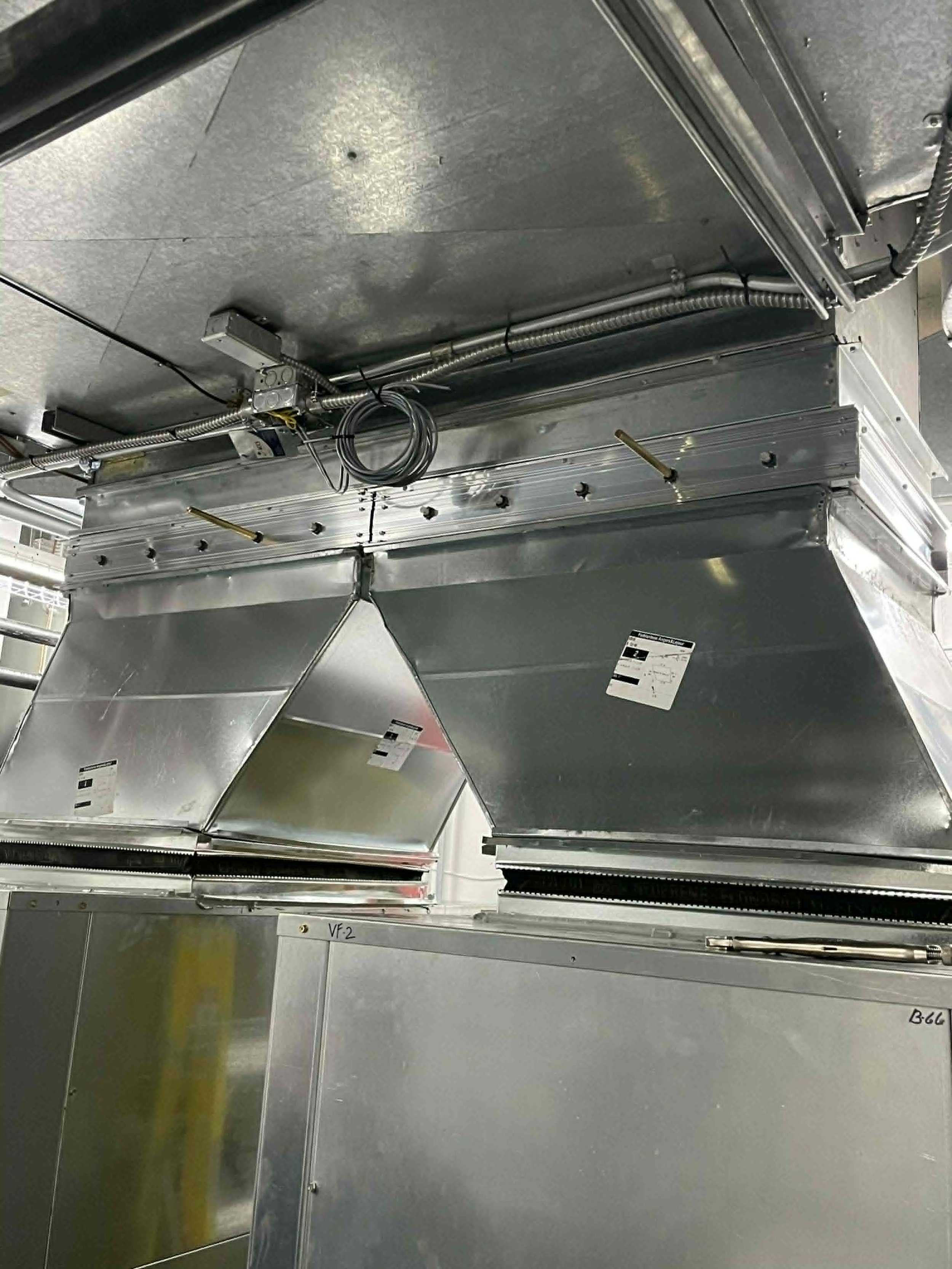 Intérieur d'une infrastructure métallique, probablement une cuisine industrielle ou une installation technique, avec des conduits, des câbles et des panneaux en métal suspendus.