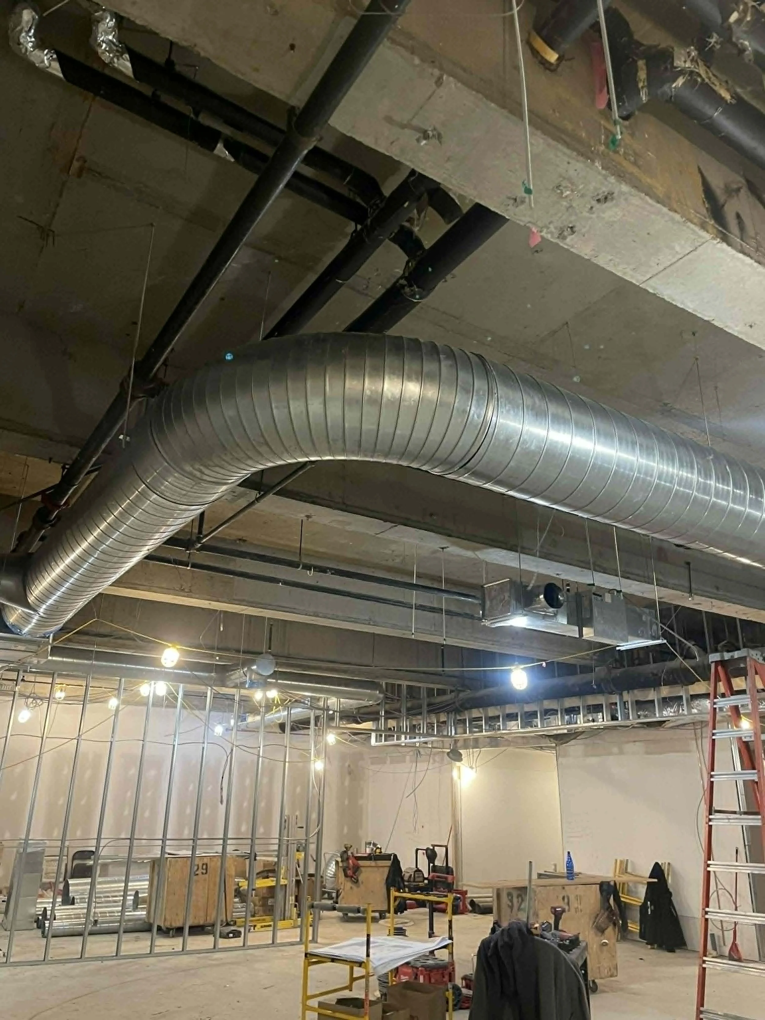 L'intérieur d'un bâtiment en construction avec des conduits d'air en métal suspendus au plafond, des outils de construction, un échafaudage rouge et des lumières brillantes installées.
