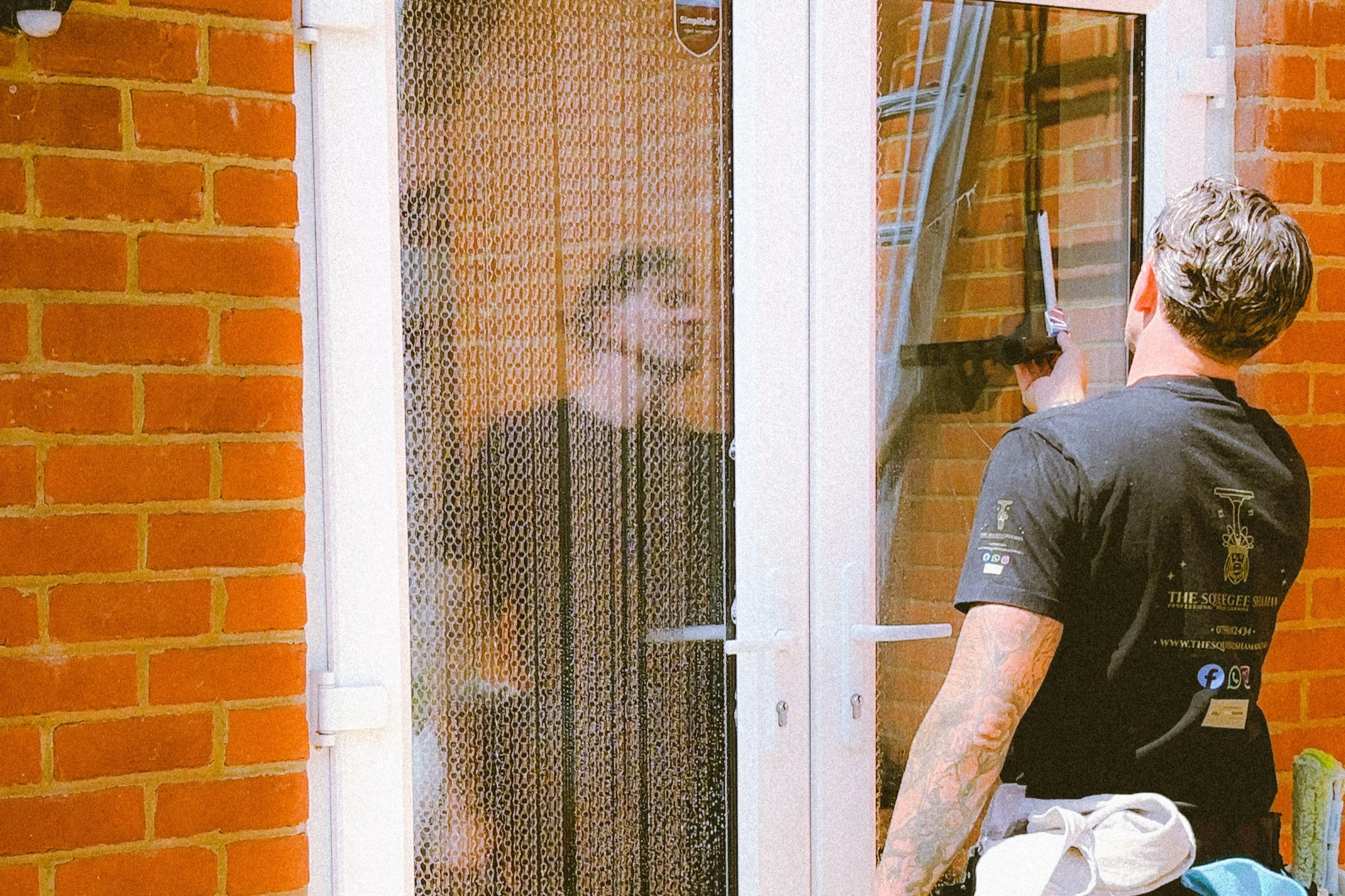 A man cleaning a door with a squeegee in East Sussex. The door has a screen with a pattern, and the background is a red brick wall.