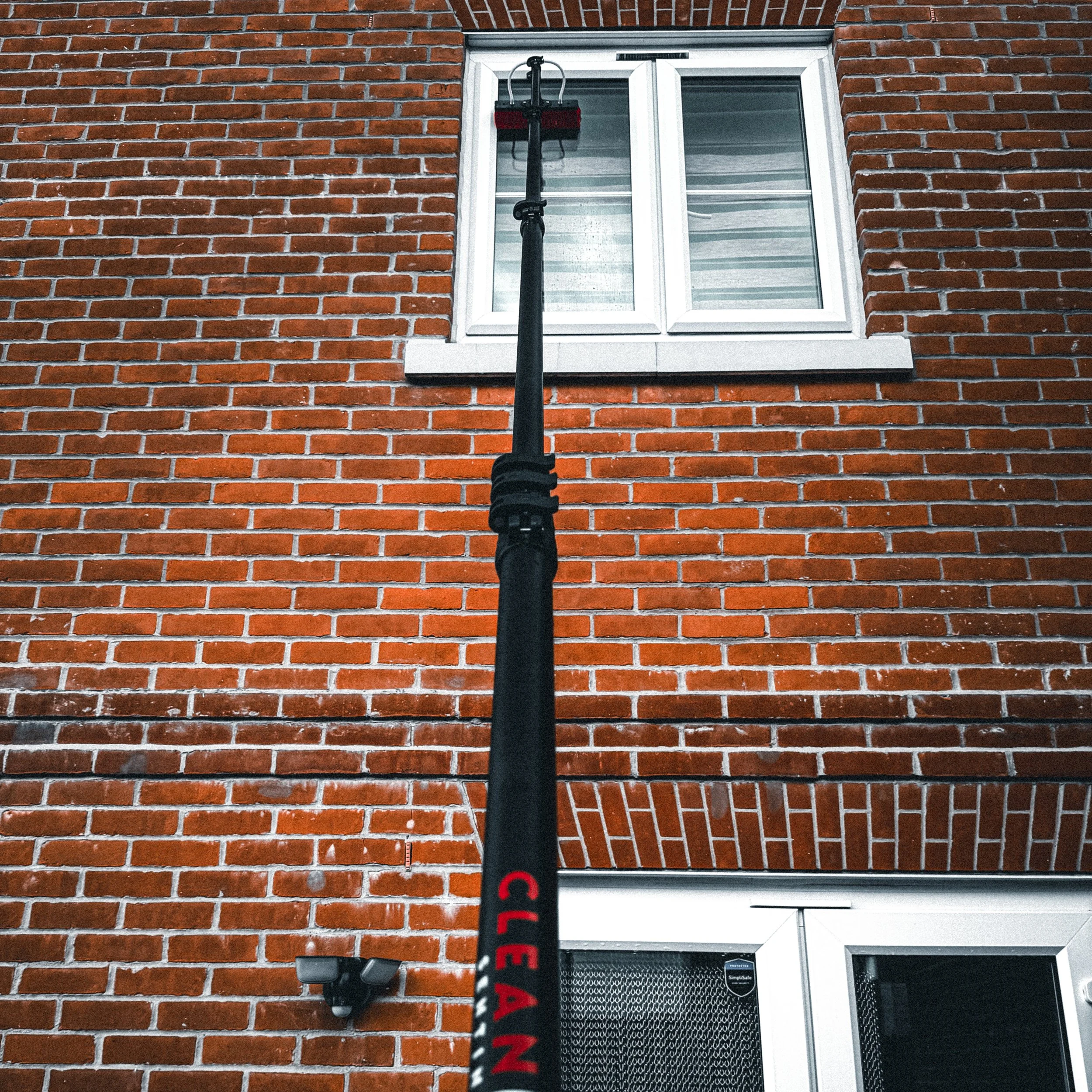 A brick building with a window and a black pole with red lettering in front of it. The pole extends upward to a platform with a handle and a cable connected to it, possibly a window cleaning tool or safety equipment.