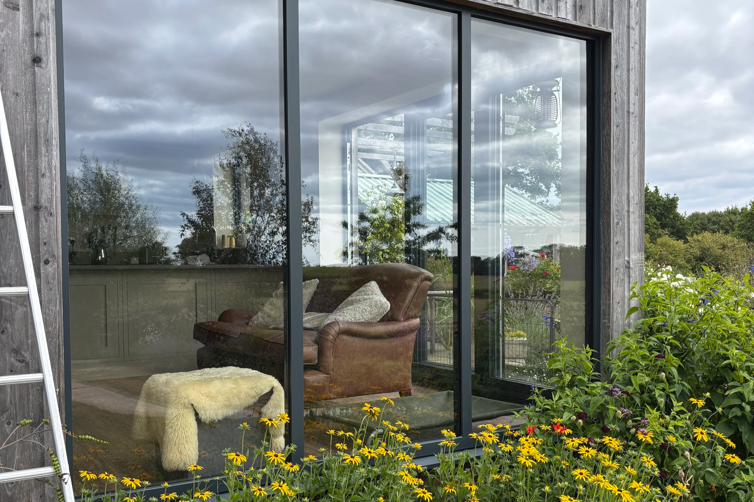 The image shows a large glass window reflecting the cloudy sky and surrounding nature. Inside, there is a cozy seating area with a vintage brown leather sofa, covered with pillows, a sheepskin rug, and some potted plants visible through the glass. Outside, there is vibrant greenery and bright yellow flowers in the garden.