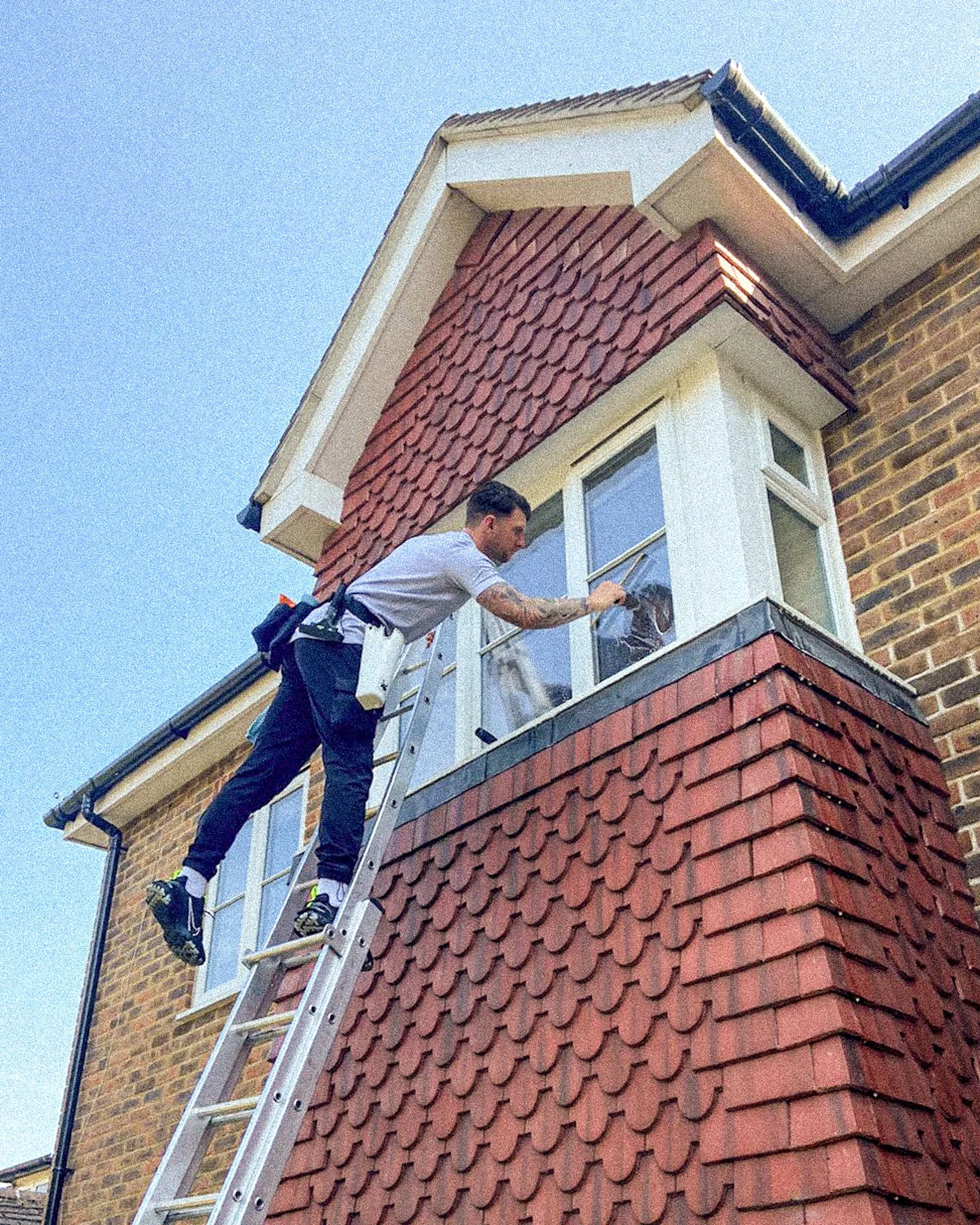 A man in a gray shirt and black pants is cleaning the exterior window of a house while standing on a ladder. The house has red shingle siding and brick walls.