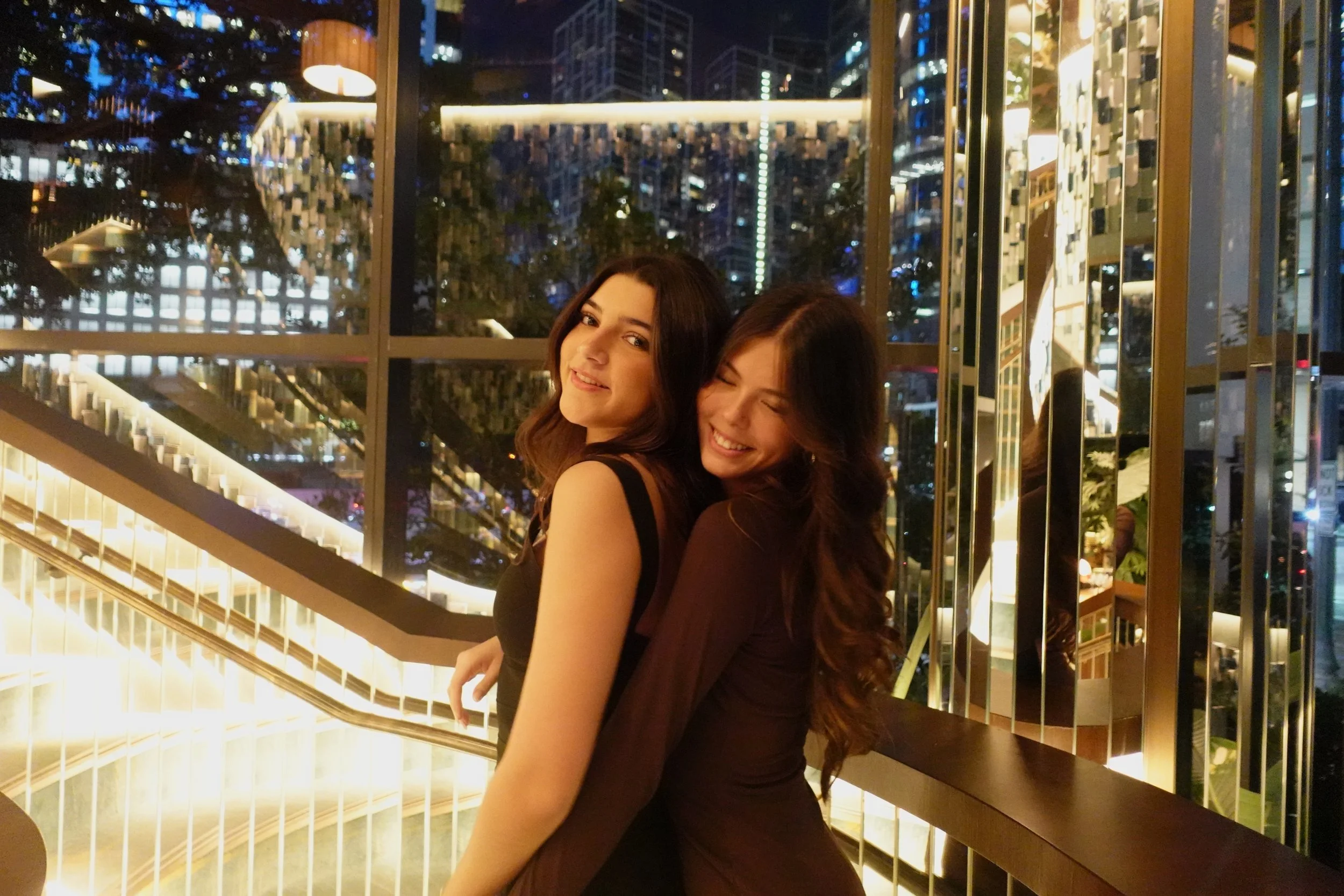 Two women laughing and smiling at night in front of a city skyscraper view with illuminated buildings and glass walls.