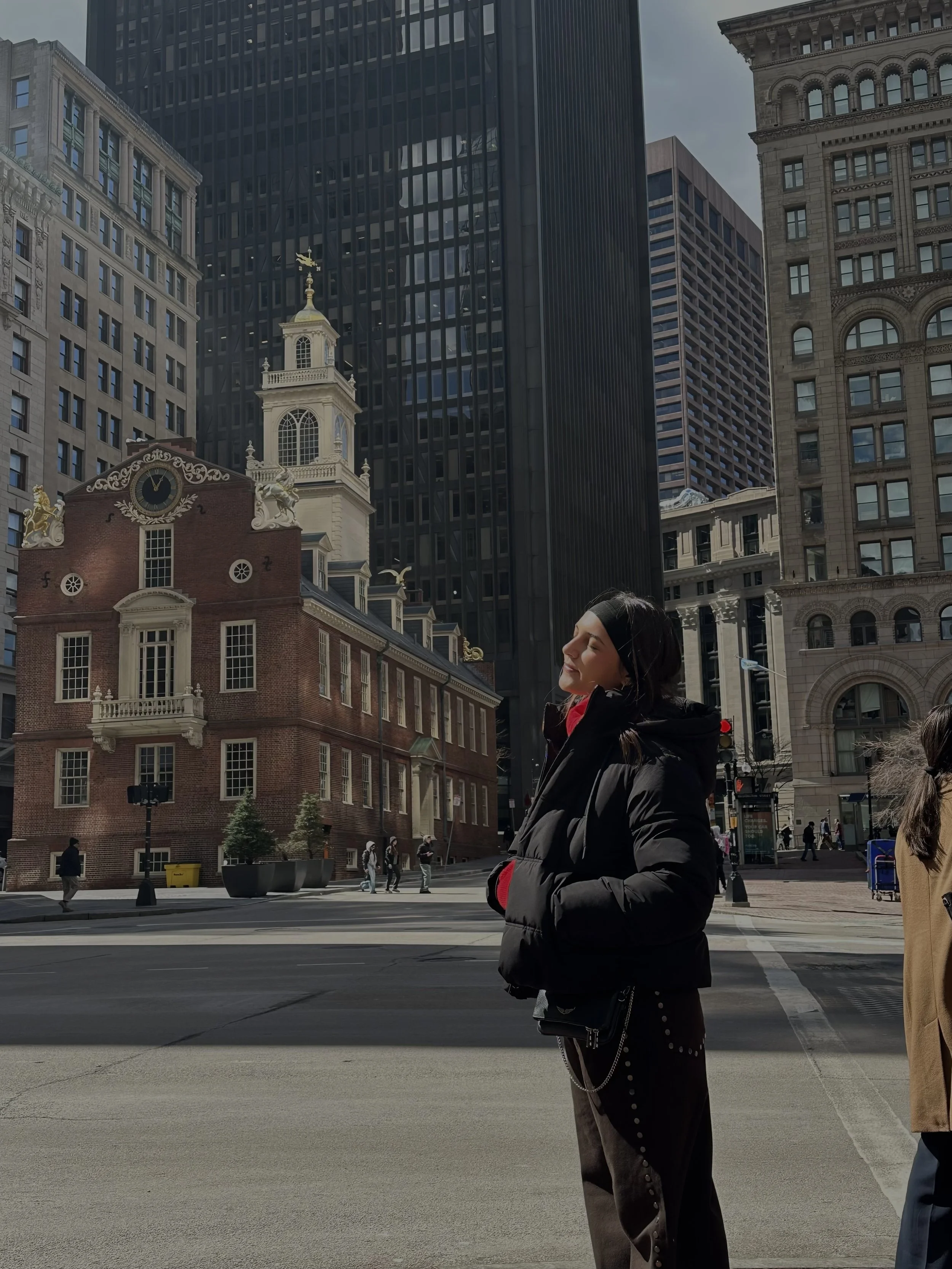 A woman with dark hair and a black headband stands on a city street during daytime, with her eyes closed and a smile. She wears a black puffy jacket and dark pants with button details. Behind her are tall buildings, including historic and modern skyscrapers, and other pedestrians walking or standing on the street.
