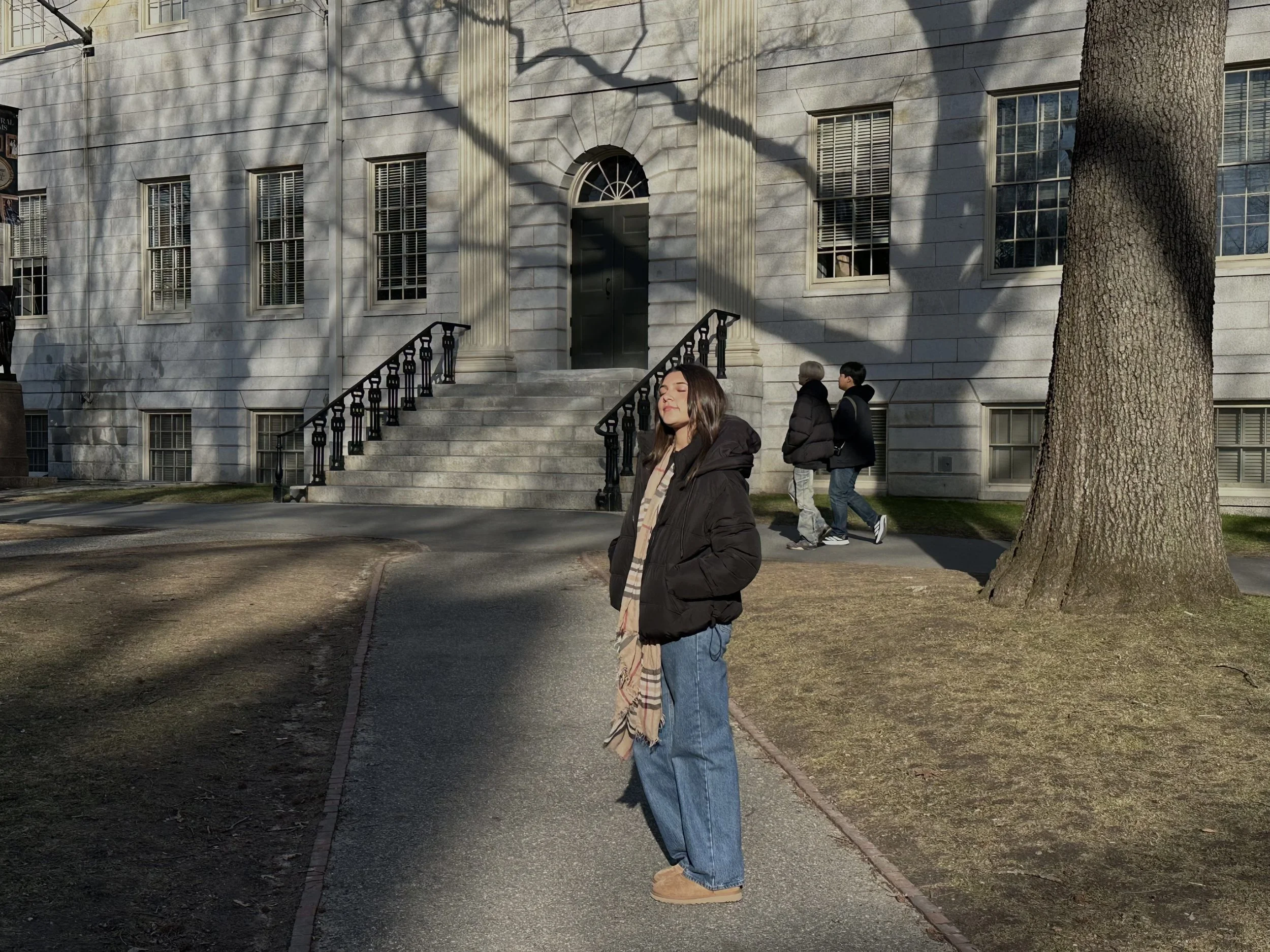 A woman with long dark hair, wearing a black jacket and a scarf, stands in front of a large white stone building with steps and black railings. Two people walk past behind her, and there is a large tree to the right, with shadows from the tree casting on the building behind her.