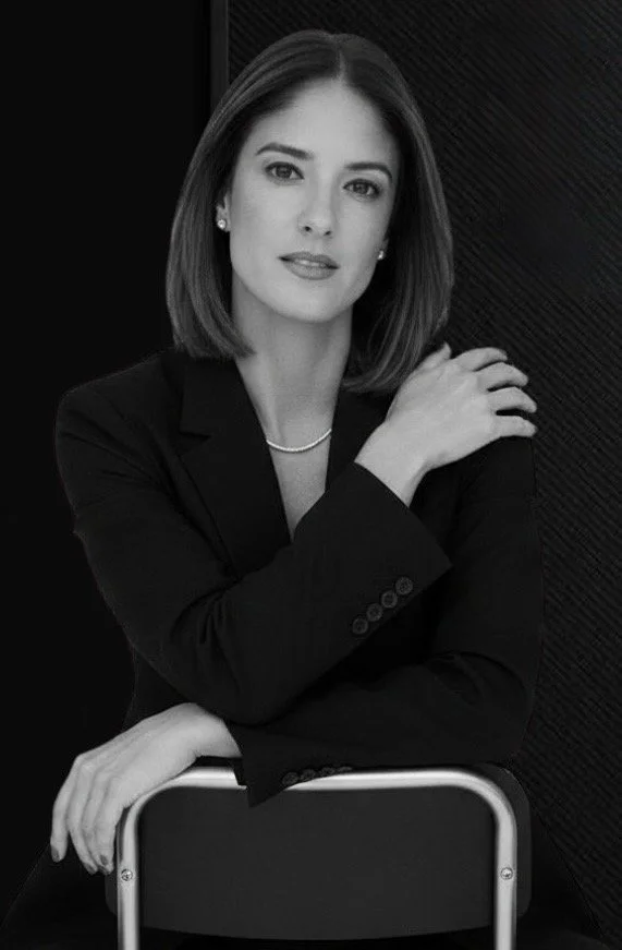 A woman with shoulder-length hair, wearing a black blazer and a pearl necklace, sitting with her right arm crossed over her chest and her left hand resting on the back of a chair, looking confidently at the camera.