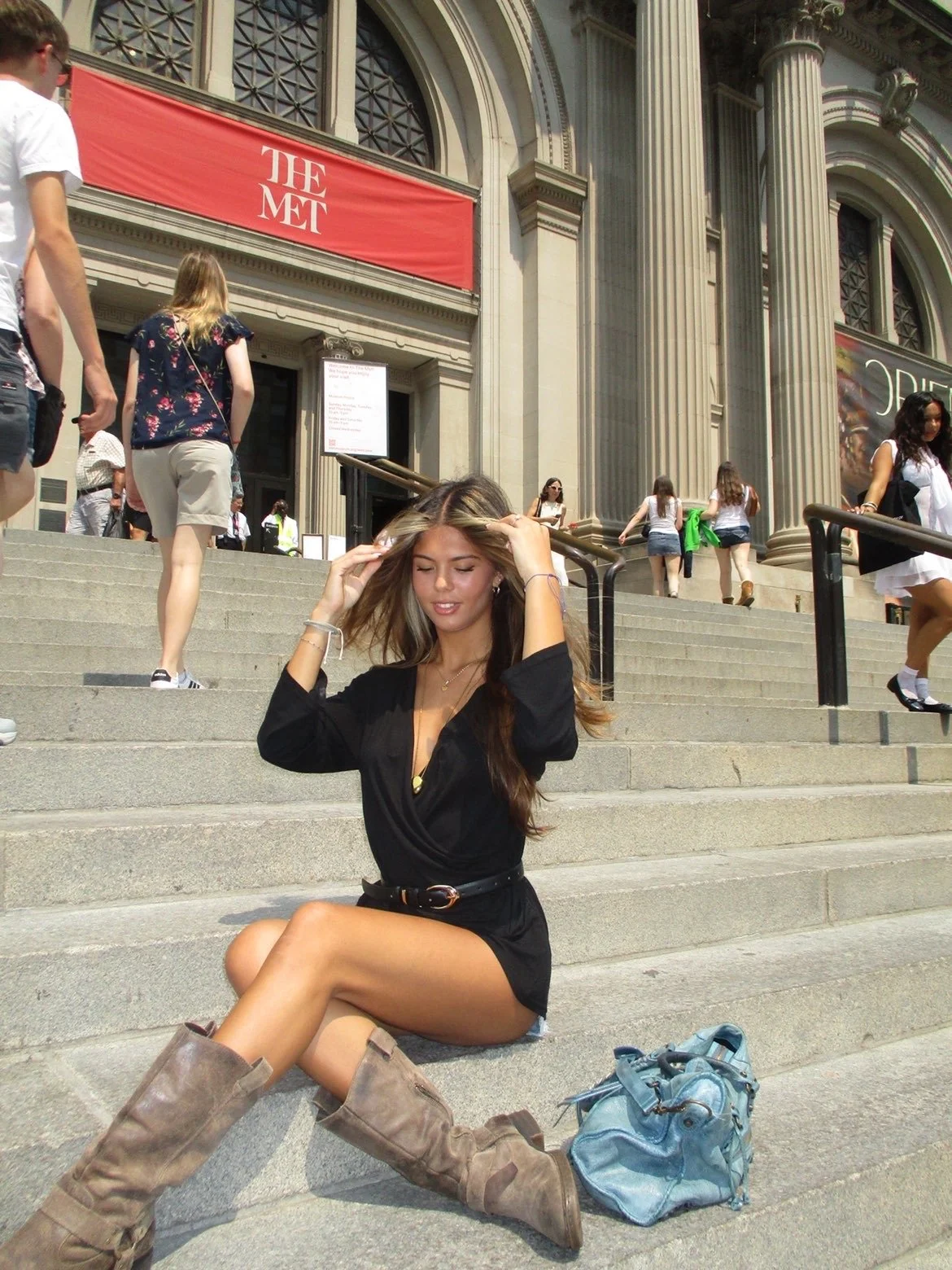 A young woman sitting on the steps in front of The Metropolitan Museum of Art in New York City, with people walking up and down the stairs behind her.