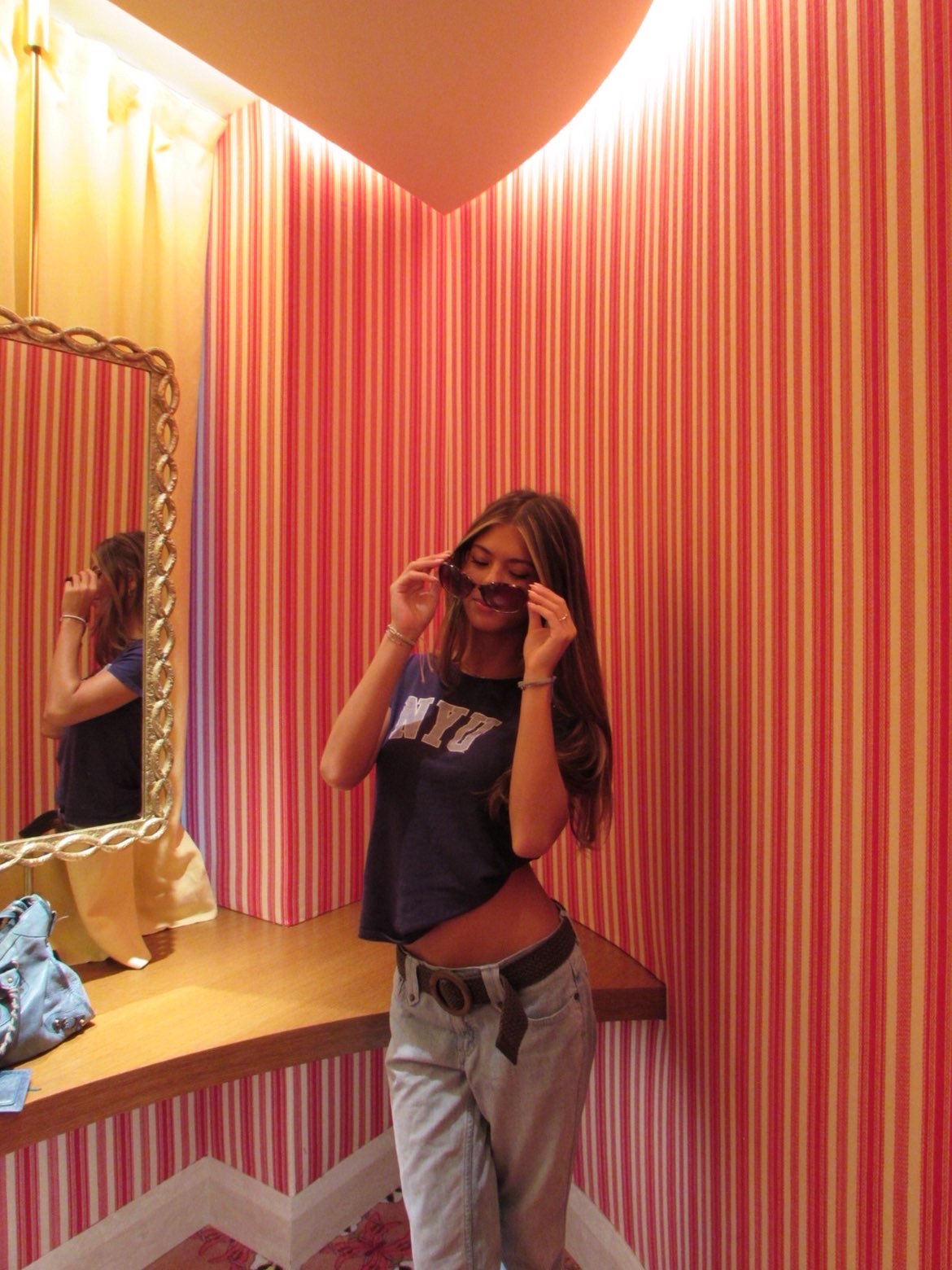 A young woman with long brown hair wearing sunglasses, a dark T-shirt, and baggy jeans poses in front of a pink and red striped wall in a dressing room, adjusting her sunglasses.