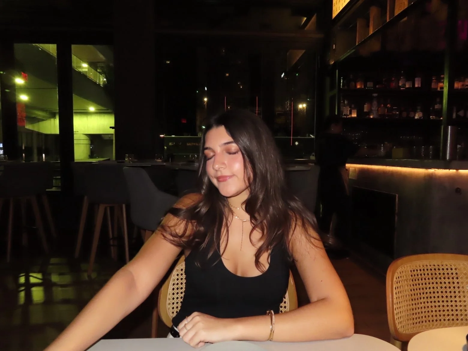 A young woman with long brown hair and fair skin, wearing a black top, sitting at a table in a dimly lit restaurant or bar at night with her eyes closed and a slight smile.