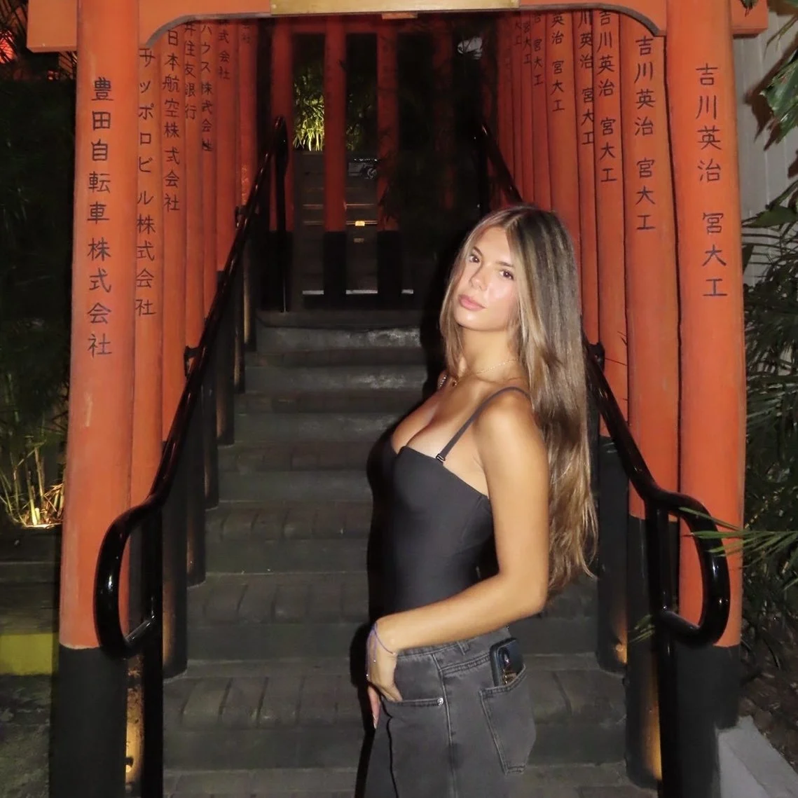 A young woman with long brown hair wearing a black spaghetti strap top and jeans, standing on a staircase with a red torii gate in the background.
