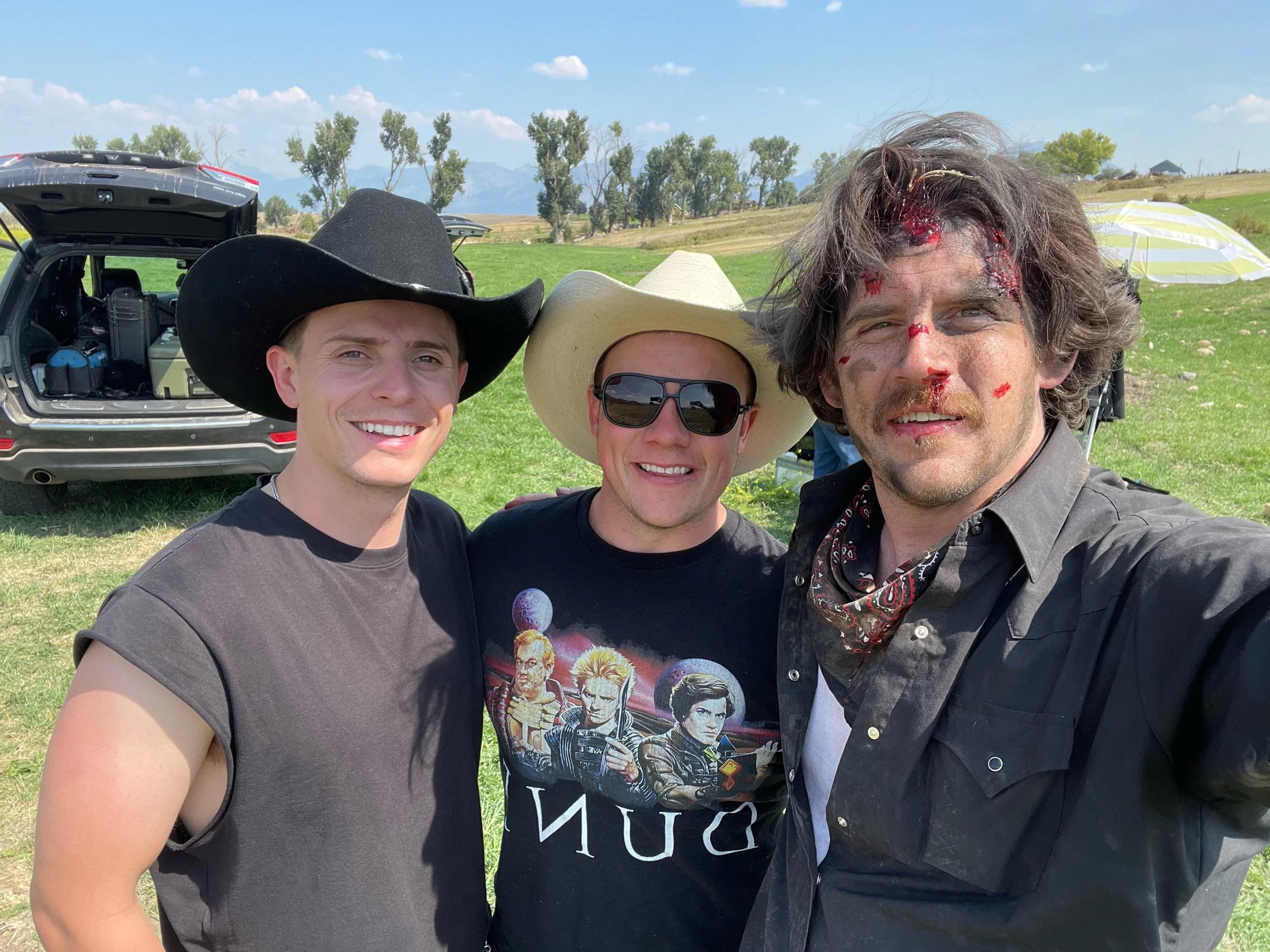 Group of three men outdoors, two wearing cowboy hats and sunglasses, one with blood and injuries on face, smiling at the camera. Open field with trees and mountains in background.