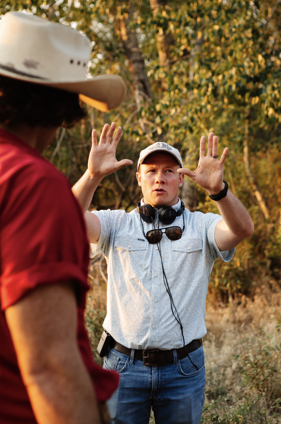 A man wearing a white cap, headphones around his neck, and a light patterned shirt, appears to be giving direction or explaining something to another person during an outdoor activity in a wooded area with autumn foliage.