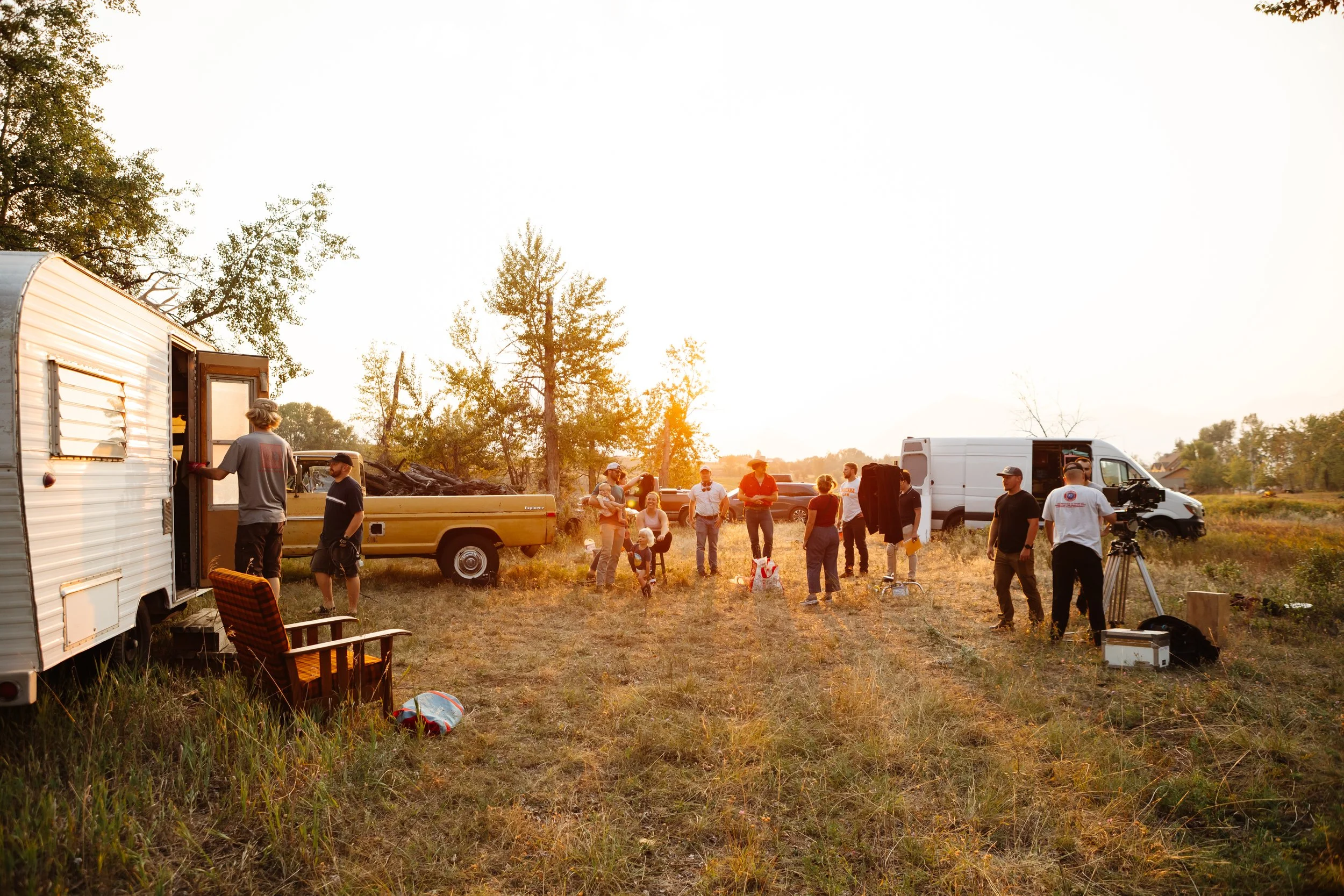 A group of people gather outside in an open field during sunset, with trees in the background and various vehicles, including a trailer, pickup trucks, and a van.