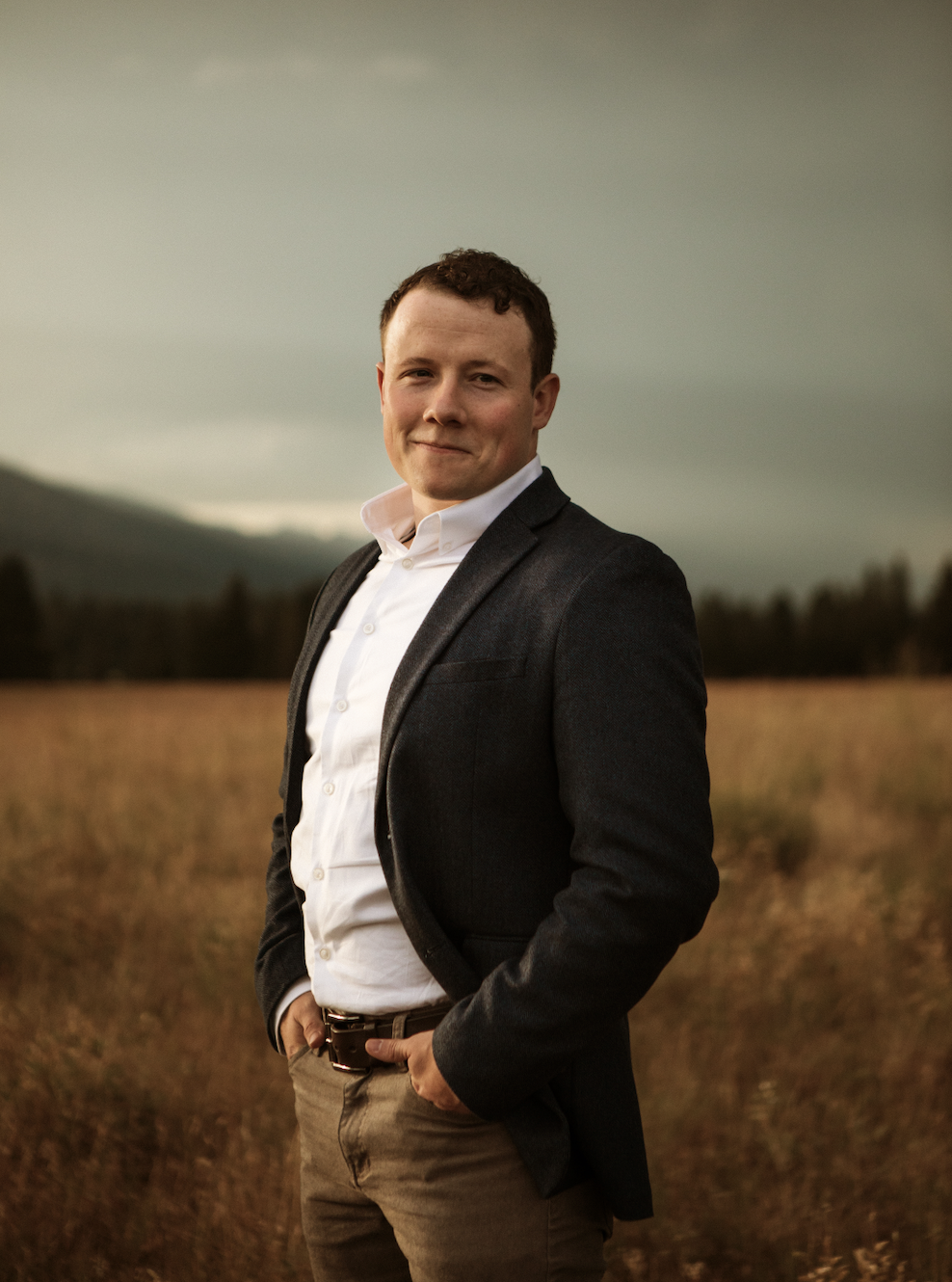 A man in business attire standing outdoors in a field with mountains in the background during sunset.