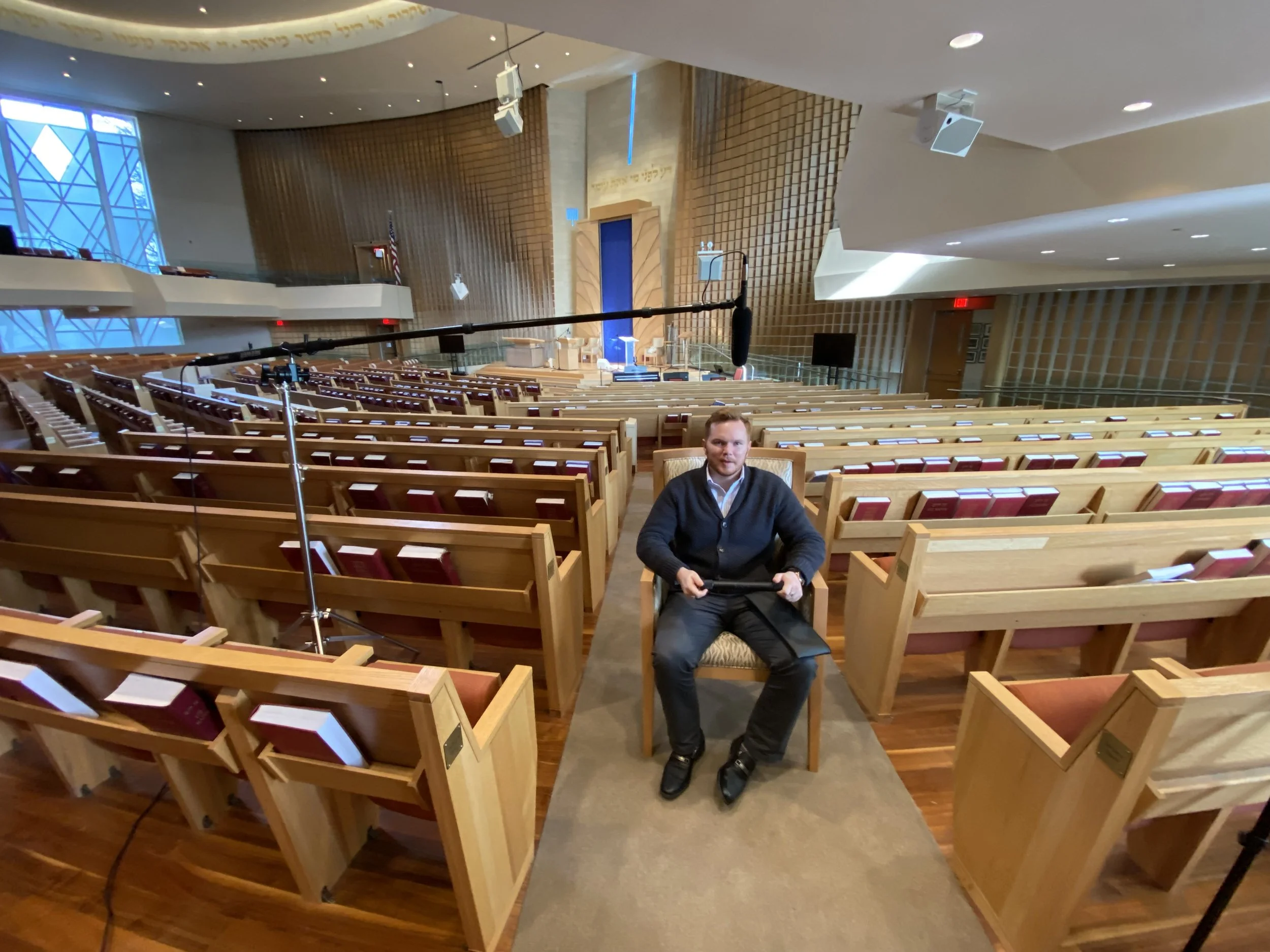 Interior of a synagogue or church with rows of wooden pews, a man sitting in a seat, and a stage area with a blue curtain and Hebrew text on the wall.