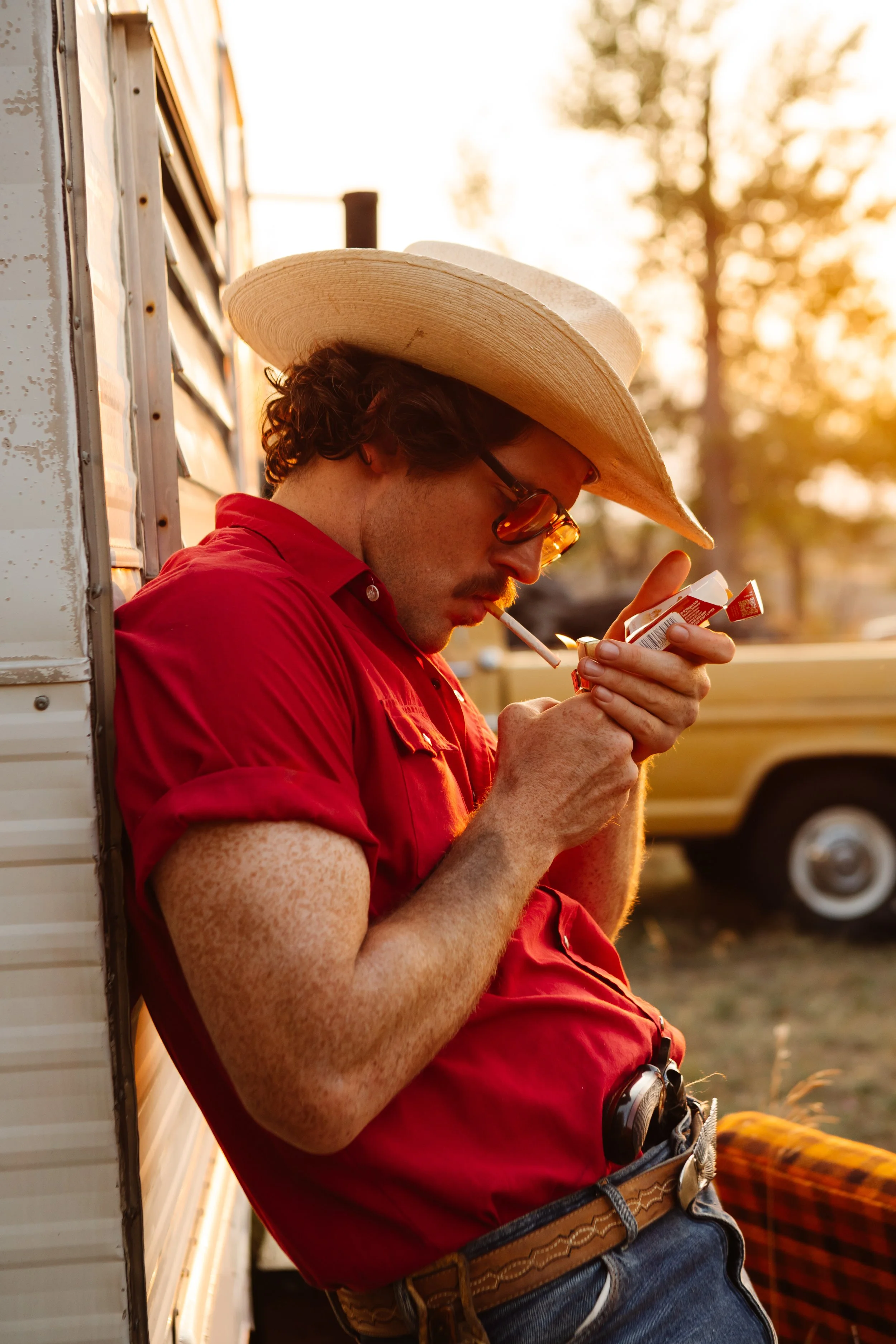 A man with curly hair and glasses, wearing a red shirt and a large straw hat, is leaning against a wall while looking at a small item in his hand. He has a cigarette in his mouth, and the scene is set outdoors during sunset with trees and a pickup truck in the background.