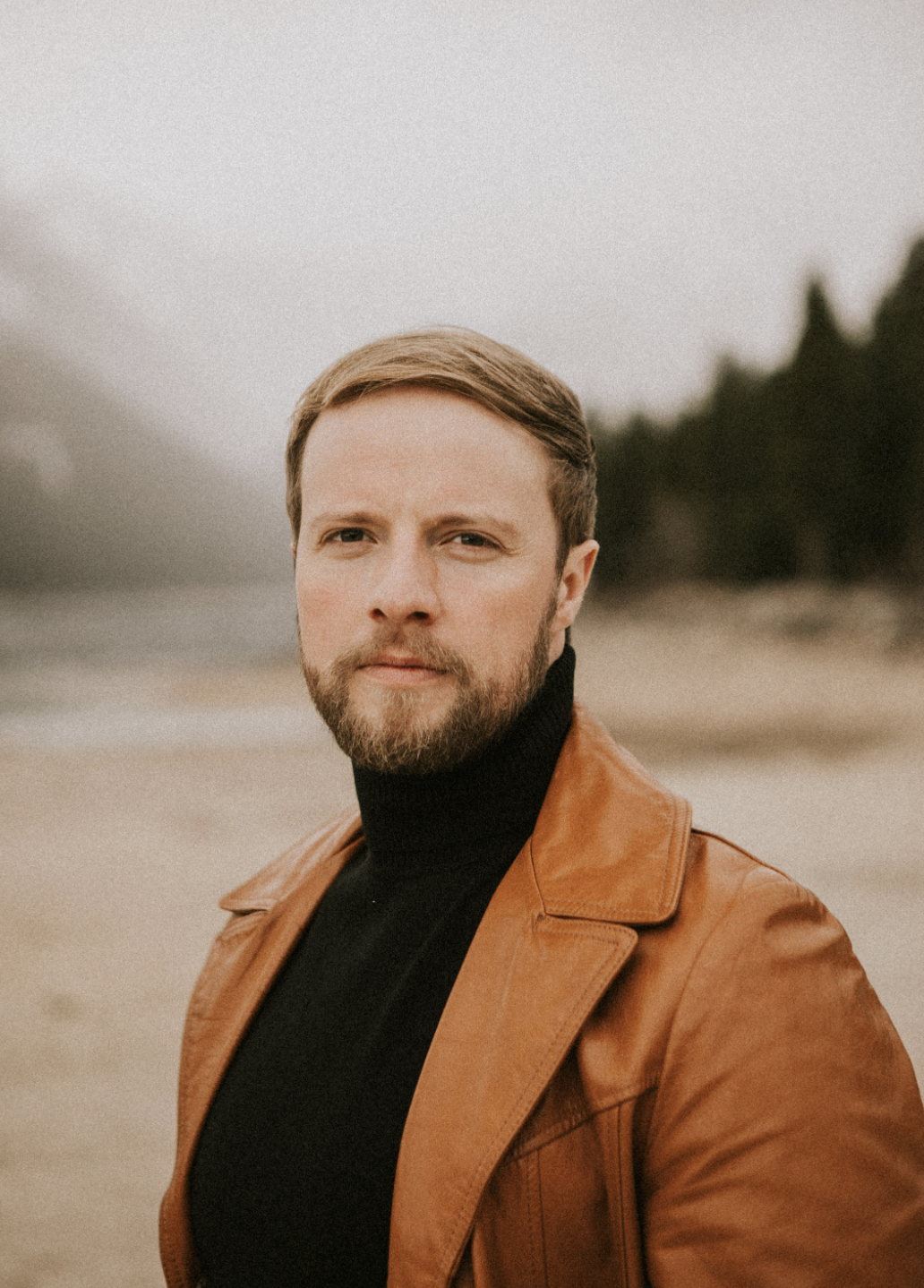 A man with a beard and short hair standing on a beach, wearing a black turtleneck and a brown leather jacket, with a blurred background of trees and cloudy sky.