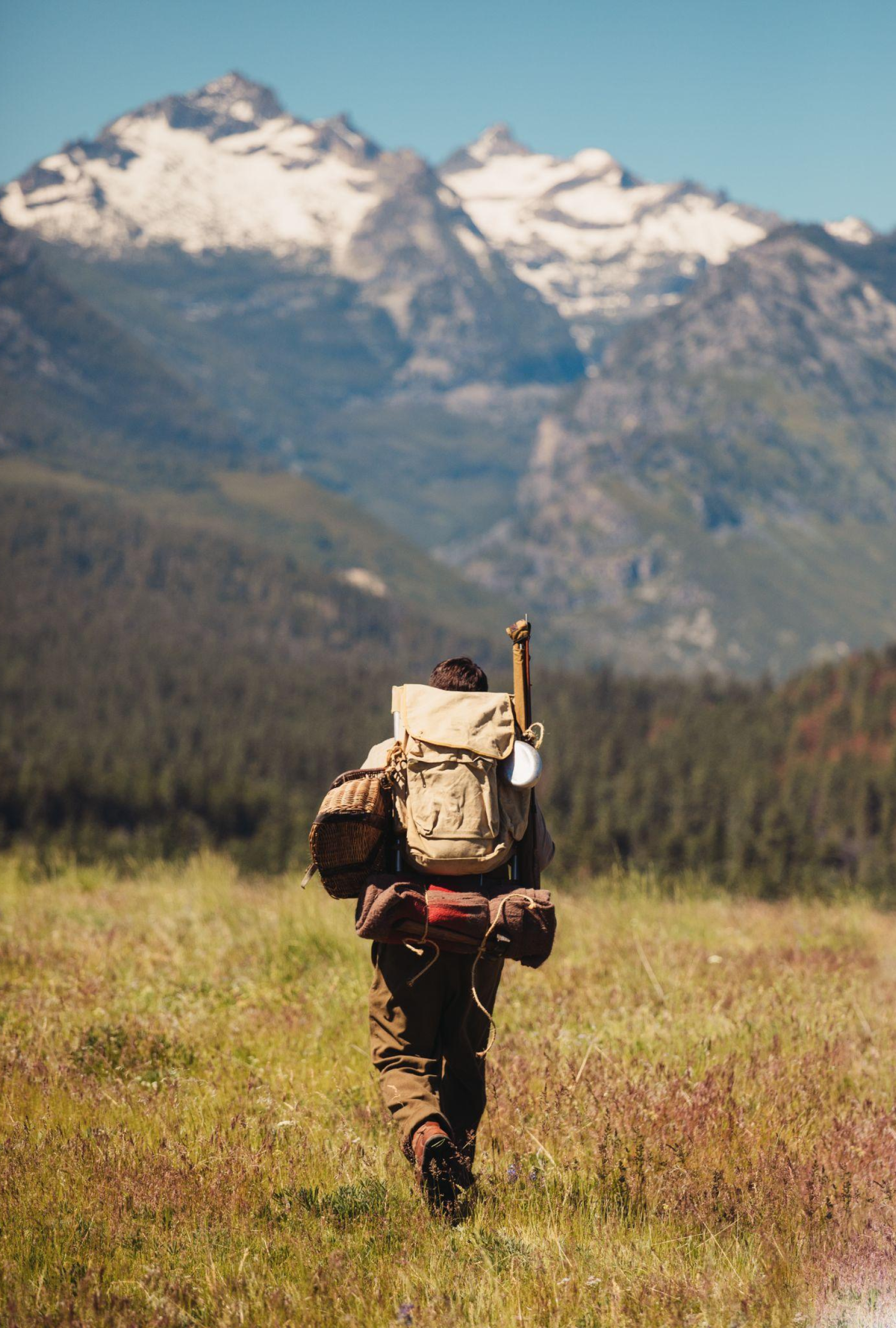 Hiker walking through a grassy field towards snow-capped mountains in the distance.