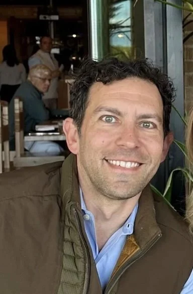 Man with curly dark hair and a blue shirt smiling at a restaurant or cafe.