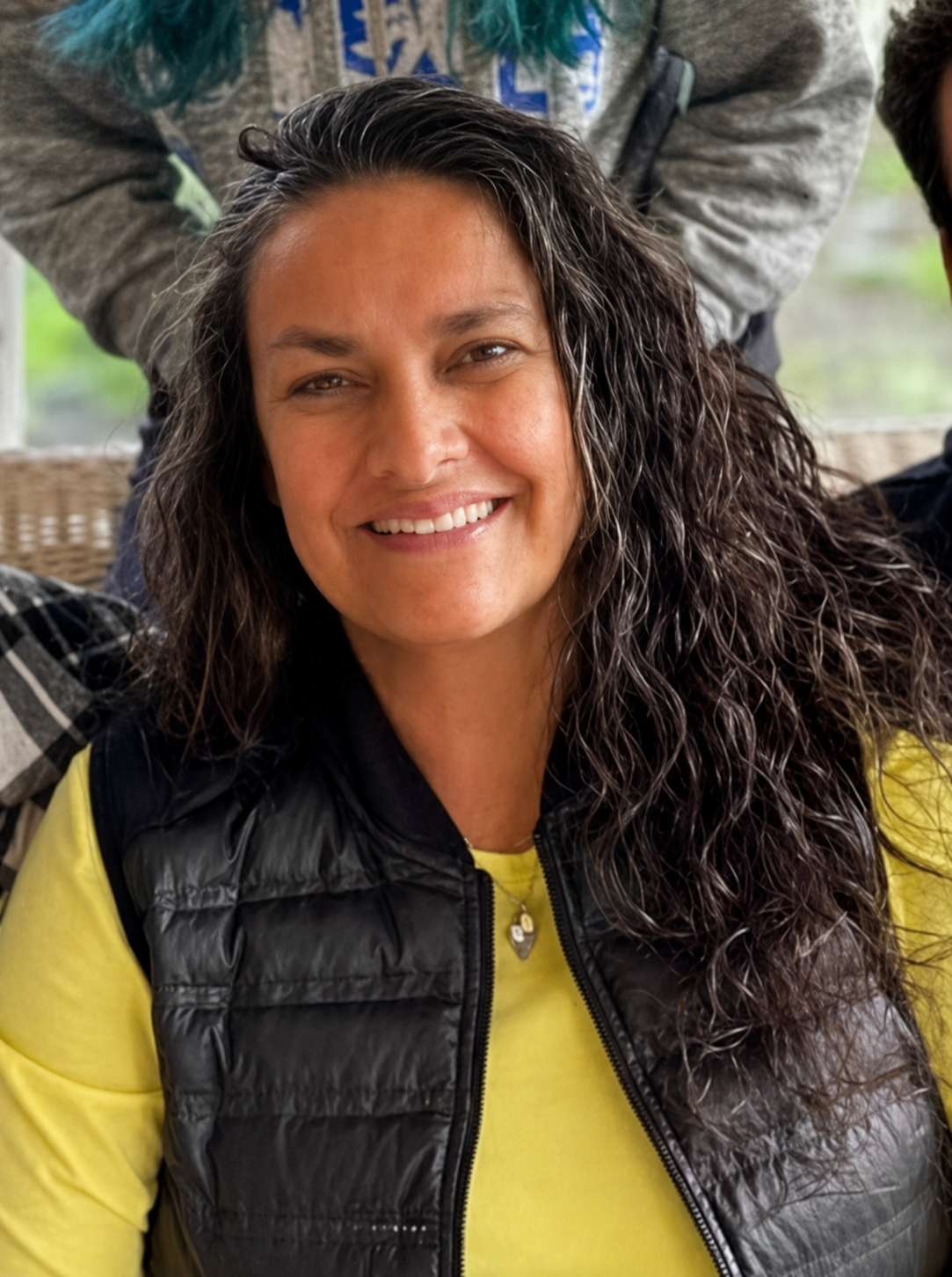 A woman with long, curly dark hair smiling at the camera, wearing a yellow shirt and a black puffer vest, with a blurred background of outdoor greenery.