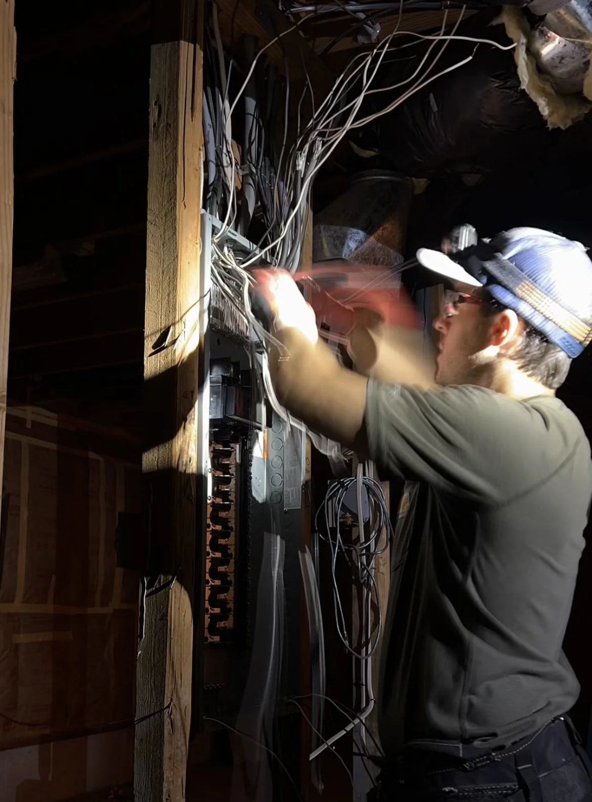 A man wearing a cap and glasses working on a wiring panel in an unfinished space.