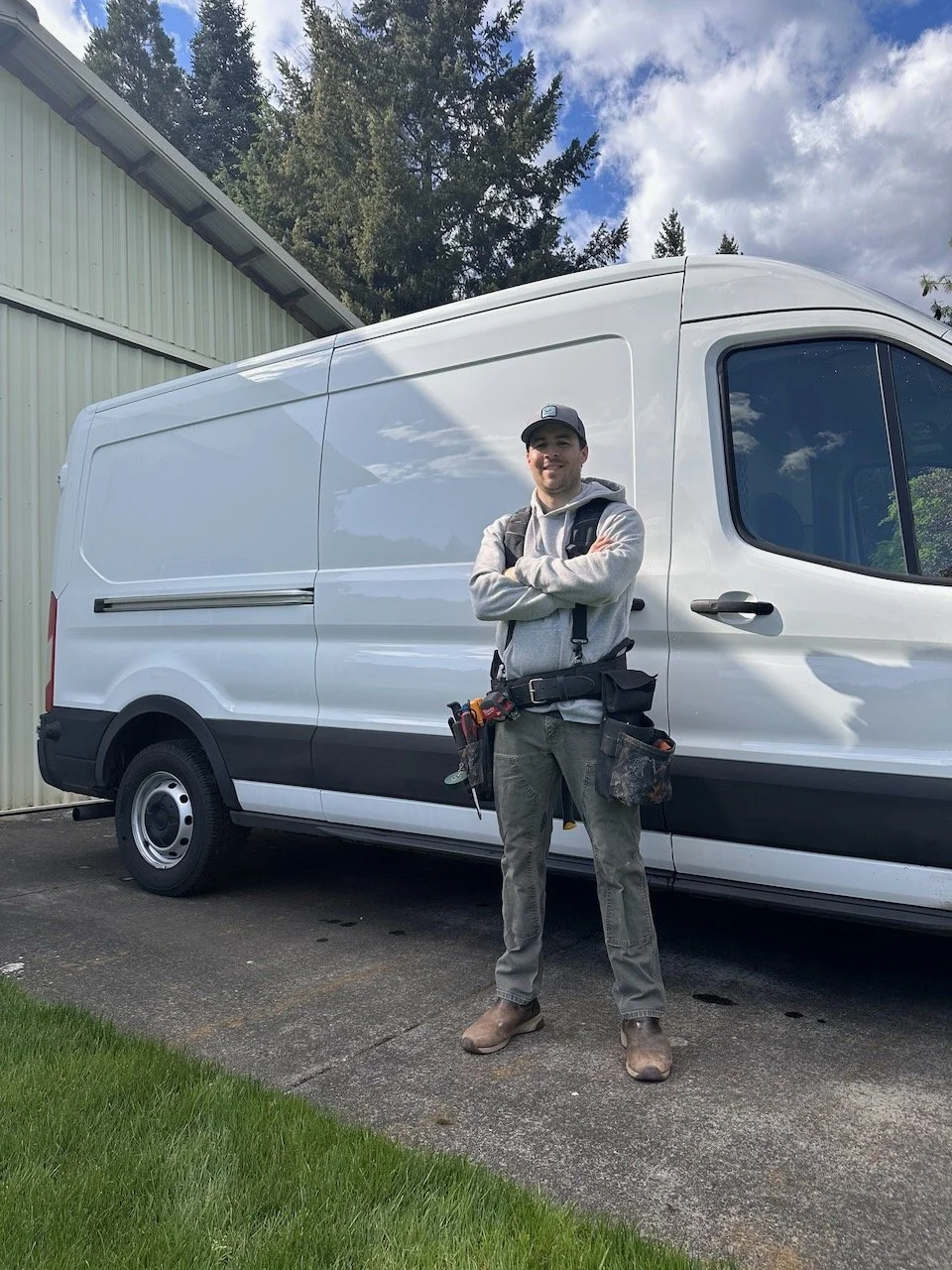 A man wearing a cap, hoodie, work pants, and boots stands with arms crossed in front of a white cargo van, with a cloudy sky and green trees in the background.