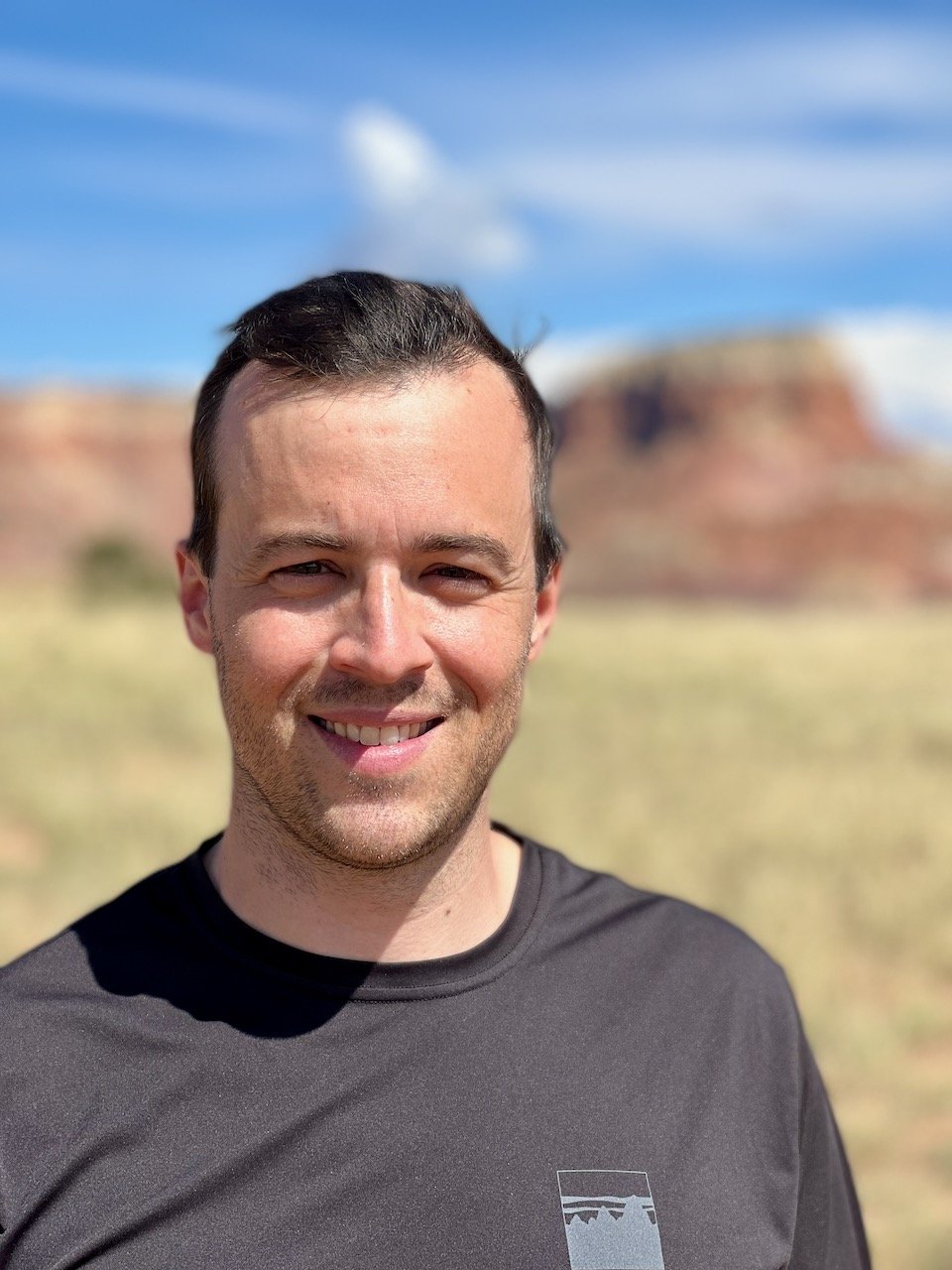 A smiling man standing outdoors with a desert landscape and red rock formations in the background under a partly cloudy sky.