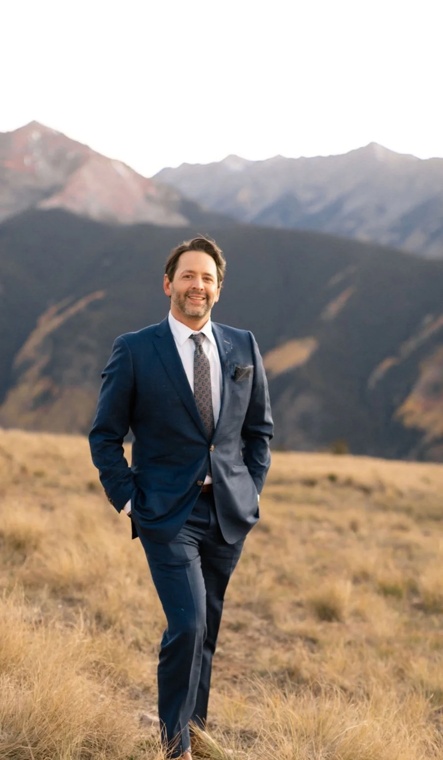 A man in a navy blue suit standing outdoors in a grassy field with mountains in the background.