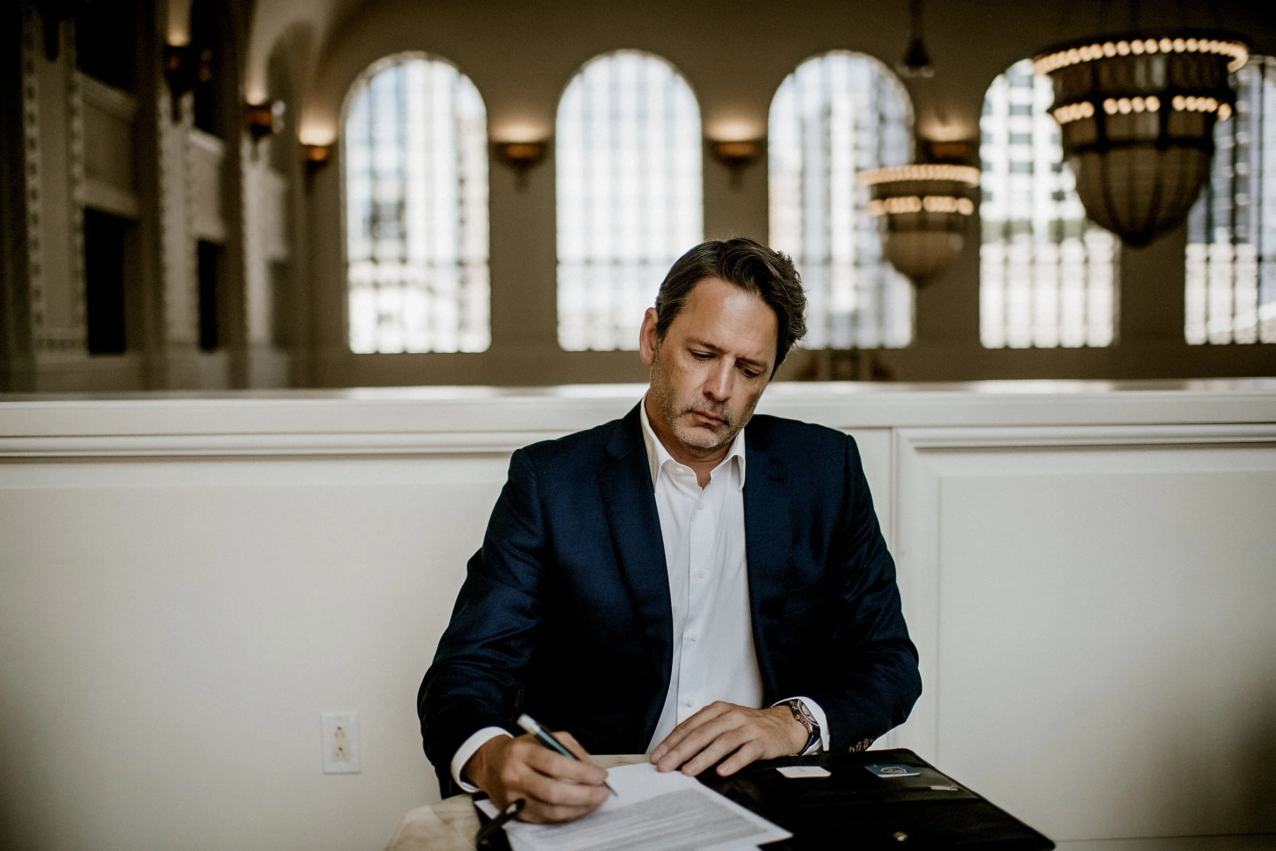 A man dressed in a dark suit and white shirt sitting at a table, writing on a piece of paper, with large windows and hanging lamps in the background.