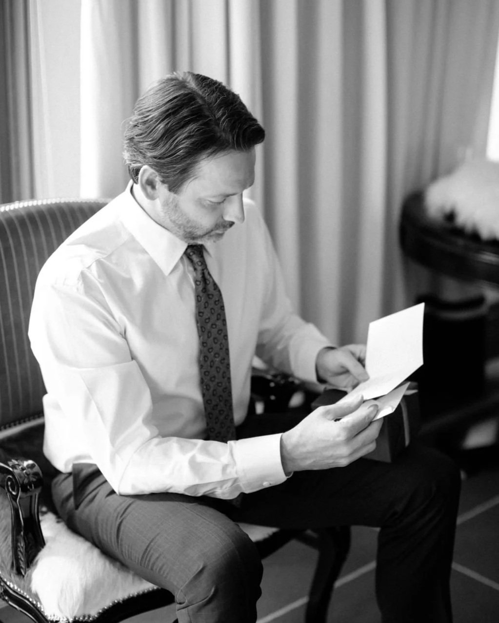 A man in formal attire, including a white shirt and tie, sitting on an ornate chair with a fur cushion, looking at a piece of paper in his hands. The background shows curtains and a side table with a closed black case.