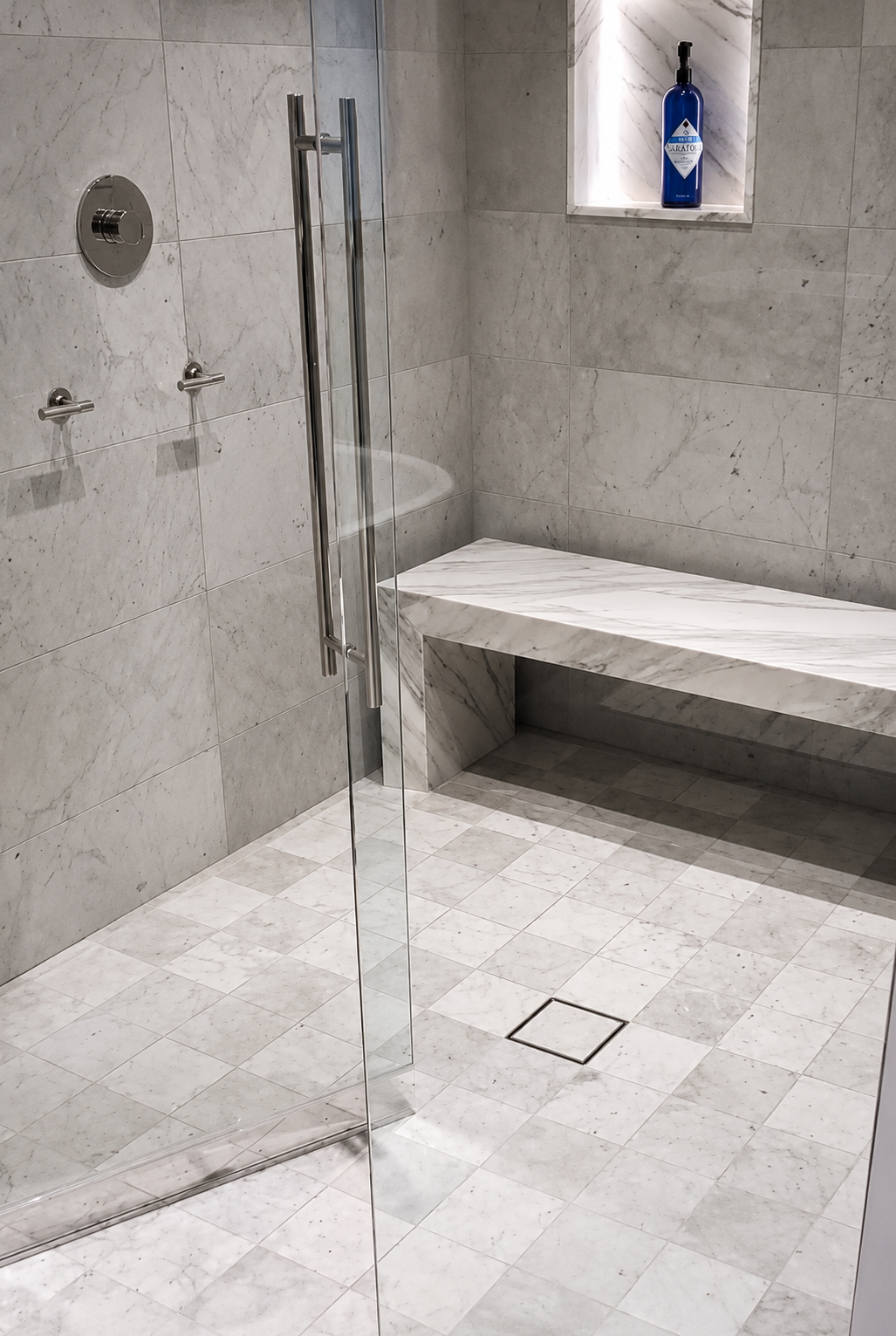A modern, minimalist shower area with marble walls and a built-in marble bench. There is a glass door with a metal handle and a niche on the wall holding a blue bottle. The floor has small marble tiles with a drain in the center.