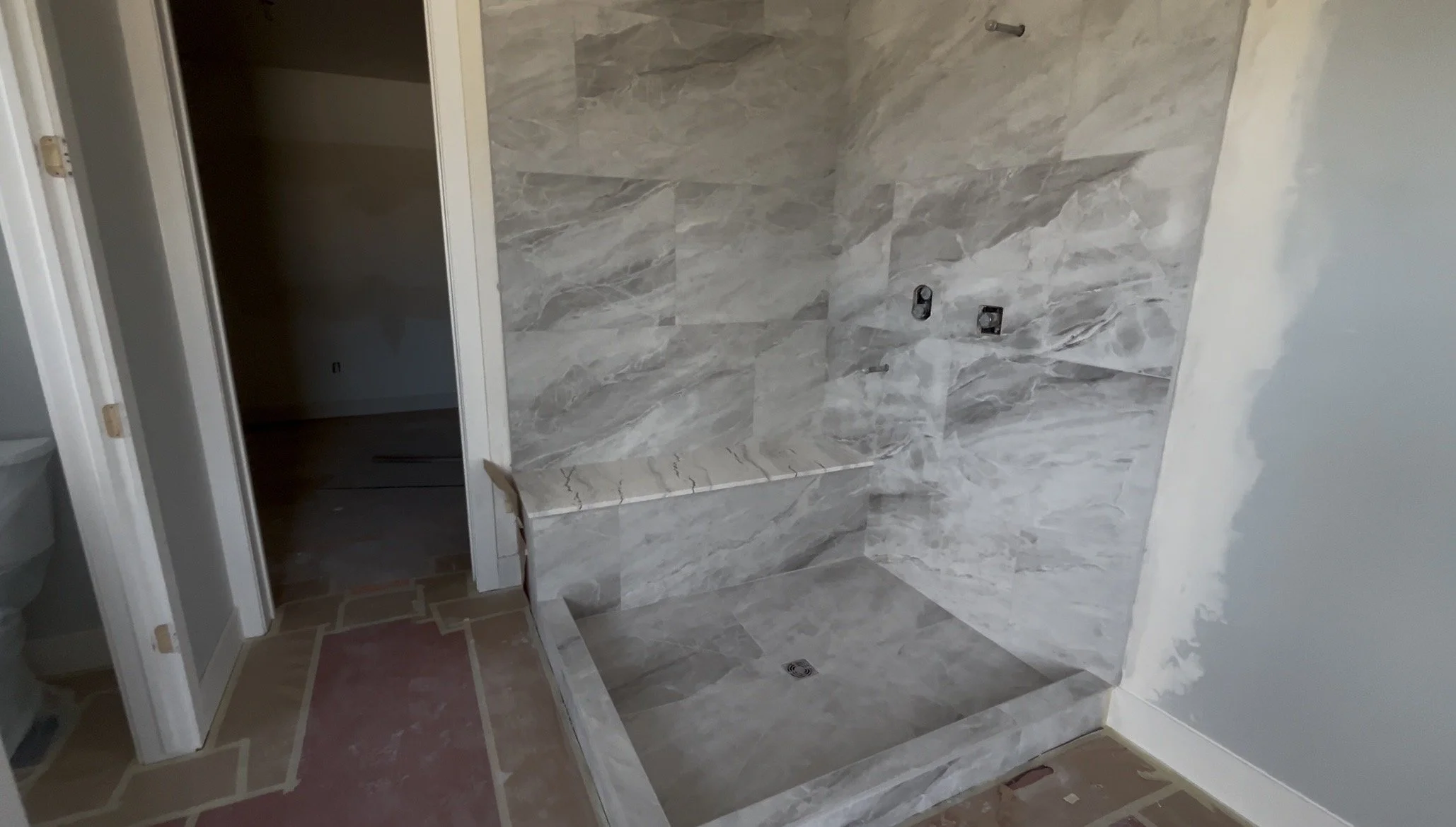 Bathroom with marble tiled shower area, unfinished wall on the right, doorway to a dark room on the left, and tiled floor.