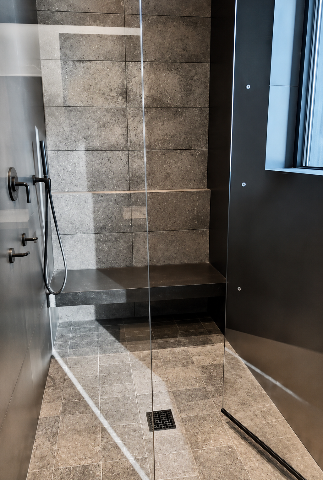 Modern, minimalistic shower with gray textured wall tiles, a glass partition, and a built-in bench. There is a window on the right wall, and a black floor drain on the tiled floor.