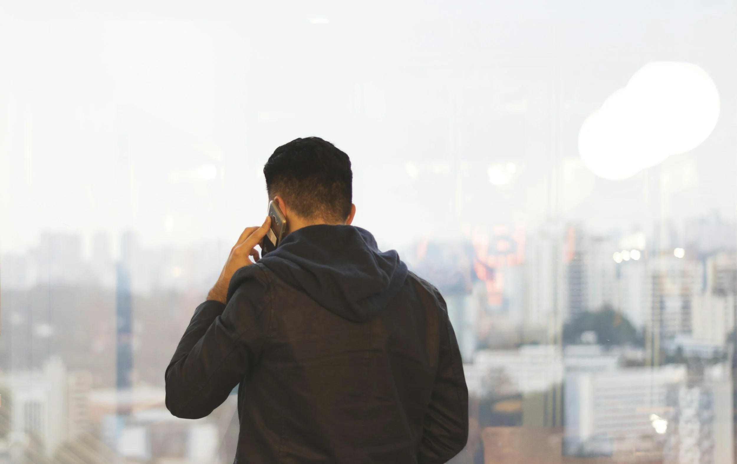 Man with short black hair, wearing a dark jacket, talking on a cell phone while looking out a large window with a cityscape in the background.