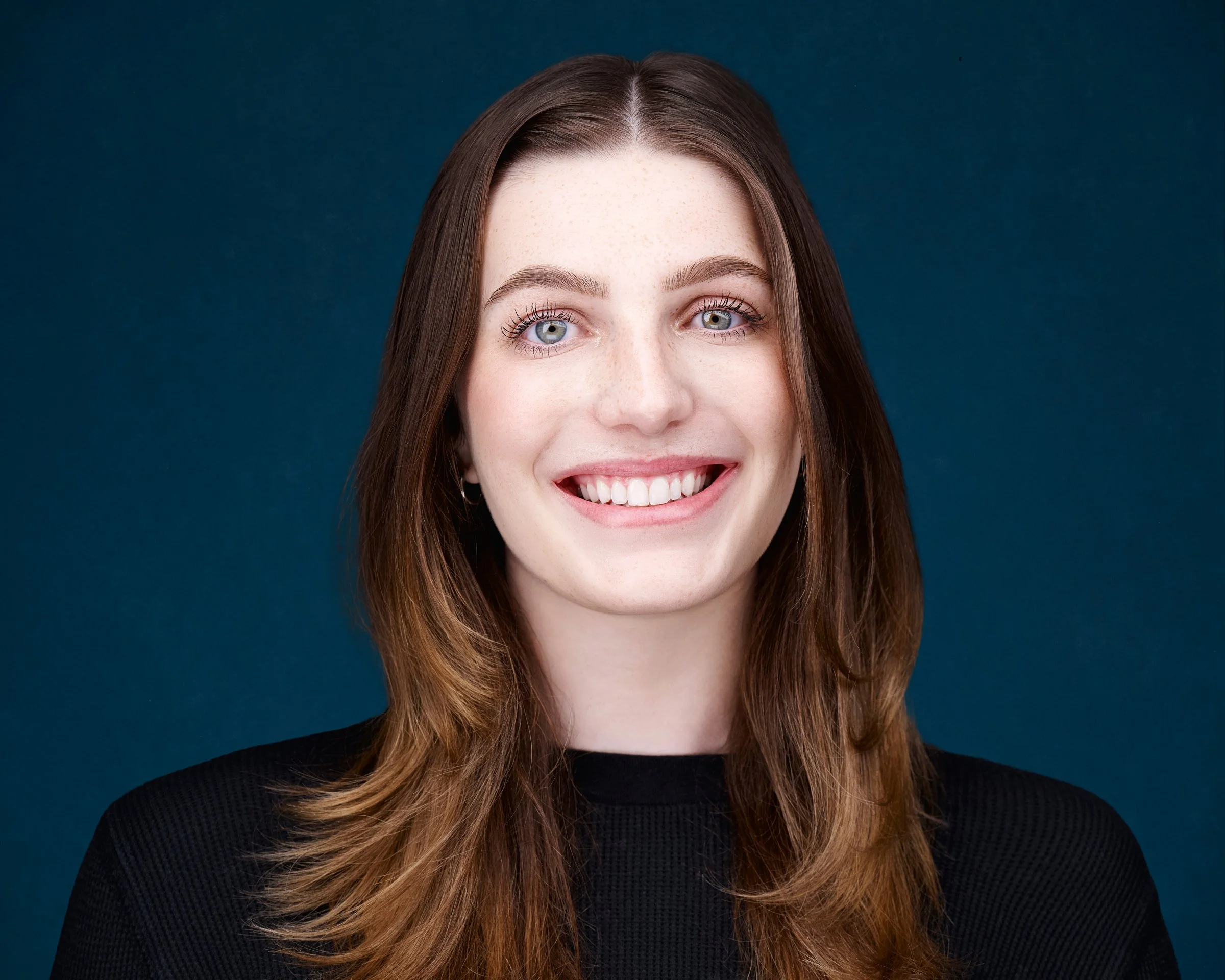 Close-up portrait of a young woman with light skin, long brown hair, blue eyes, and a bright smile, wearing a black top against a dark blue background.