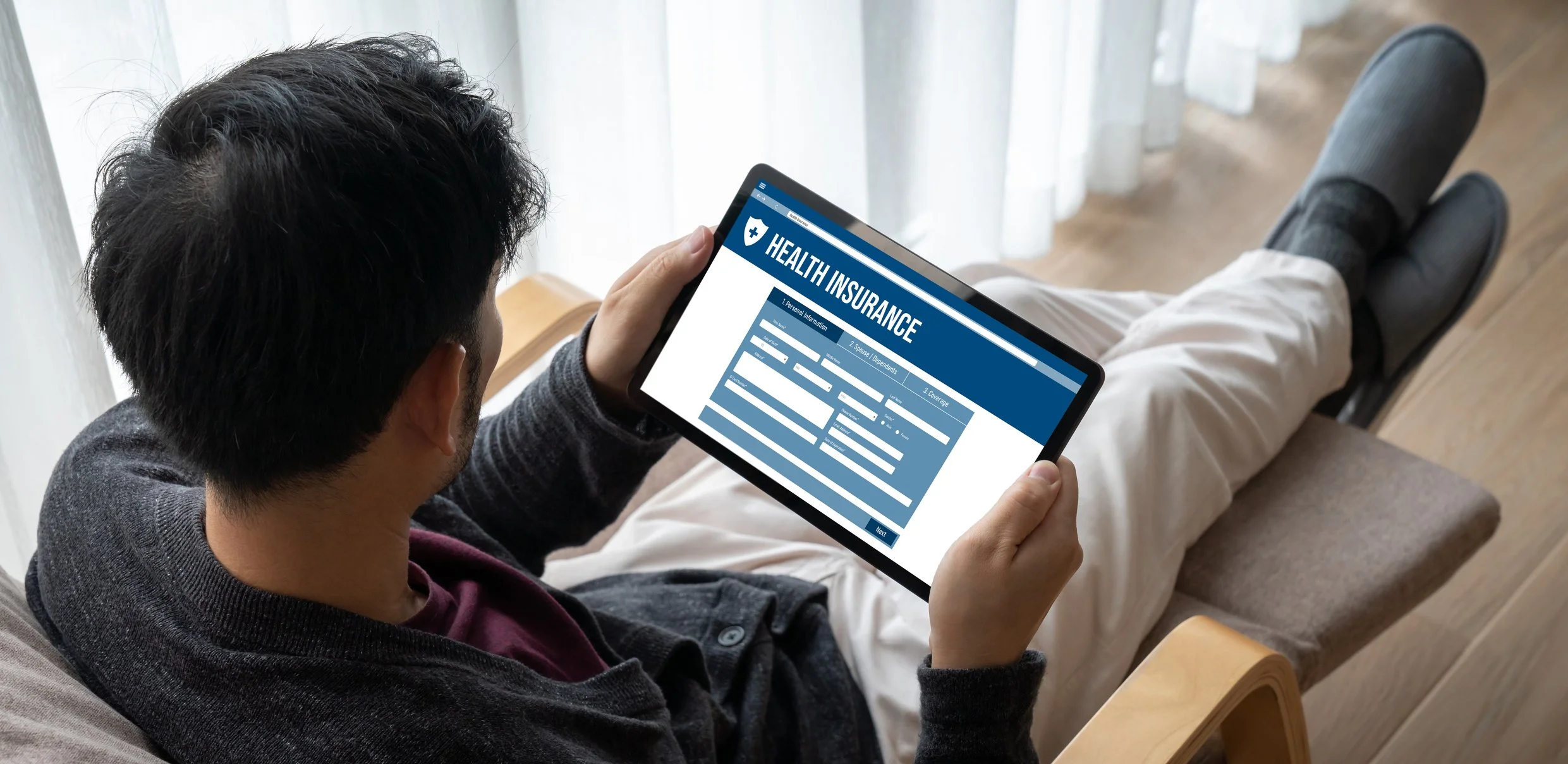 Person holding a tablet displaying a health insurance form, seated on a beige chair with crossed legs, near a window with sheer curtains.