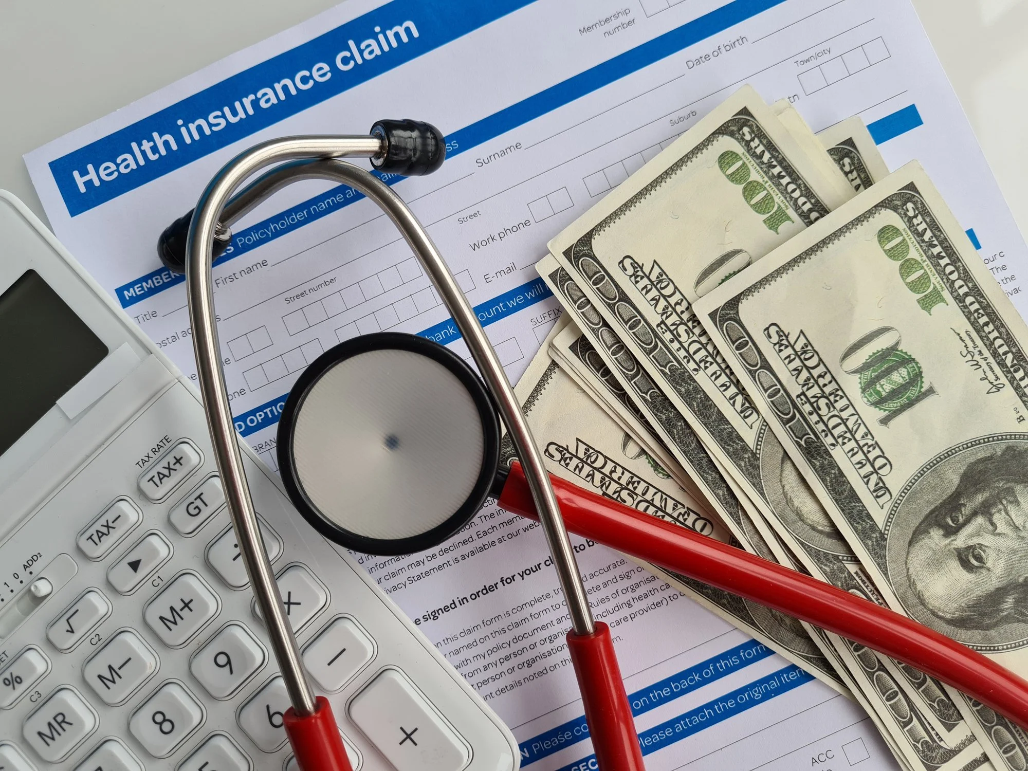 An insurance claim form with a stethoscope, several hundred dollar bills, a red pen, a calculator, and some paperwork on a white surface.