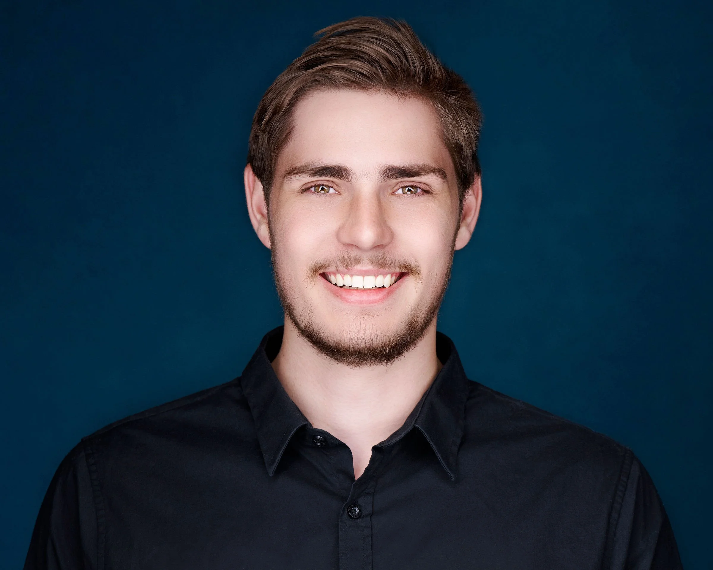 Close-up portrait of a young man with a beard and light brown hair, smiling against a dark blue background.