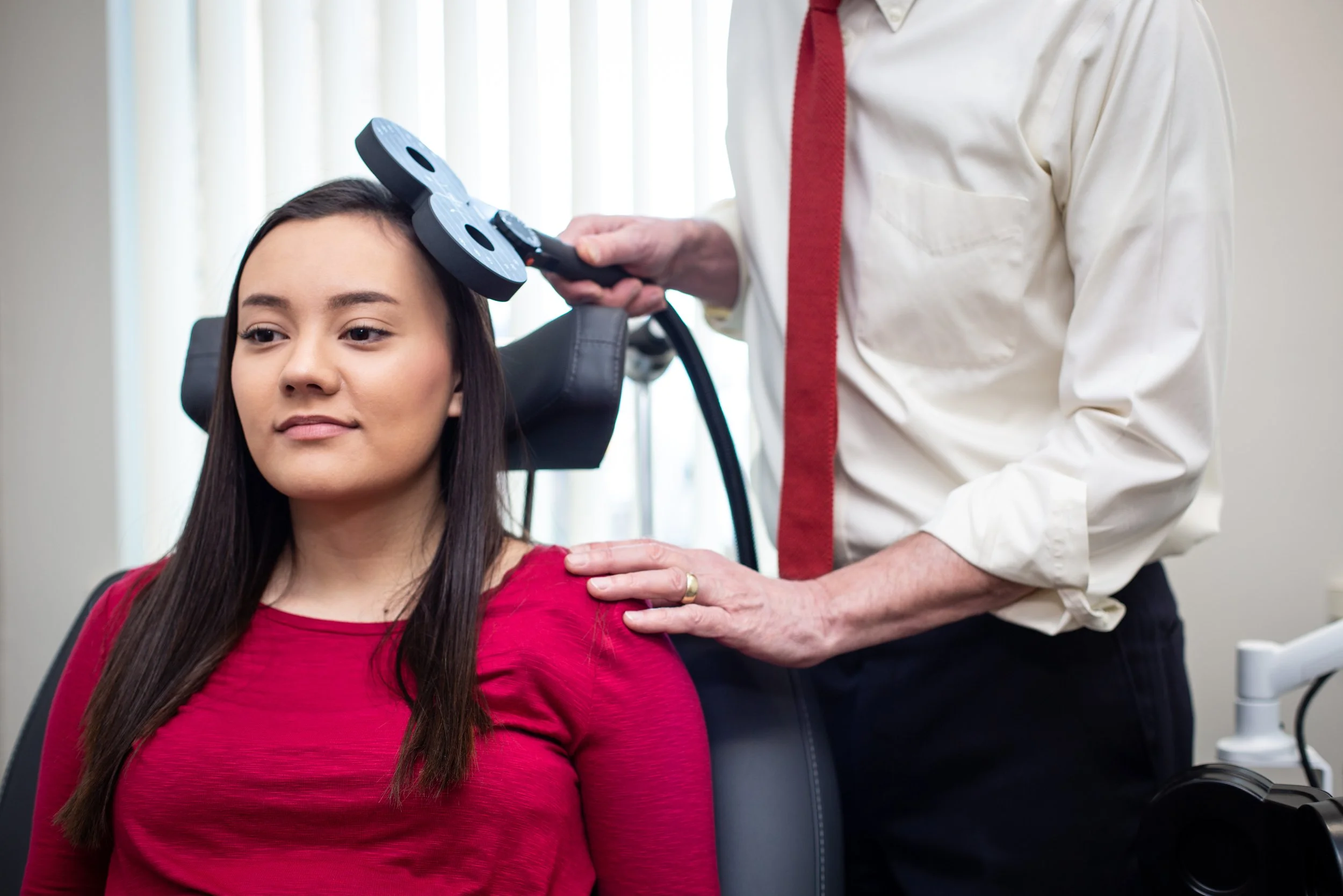 A woman with long dark hair wearing a red top is sitting in a medical chair, receiving a vaccination or injection in her upper arm from a healthcare professional wearing a white shirt and red tie, in a clinical setting.