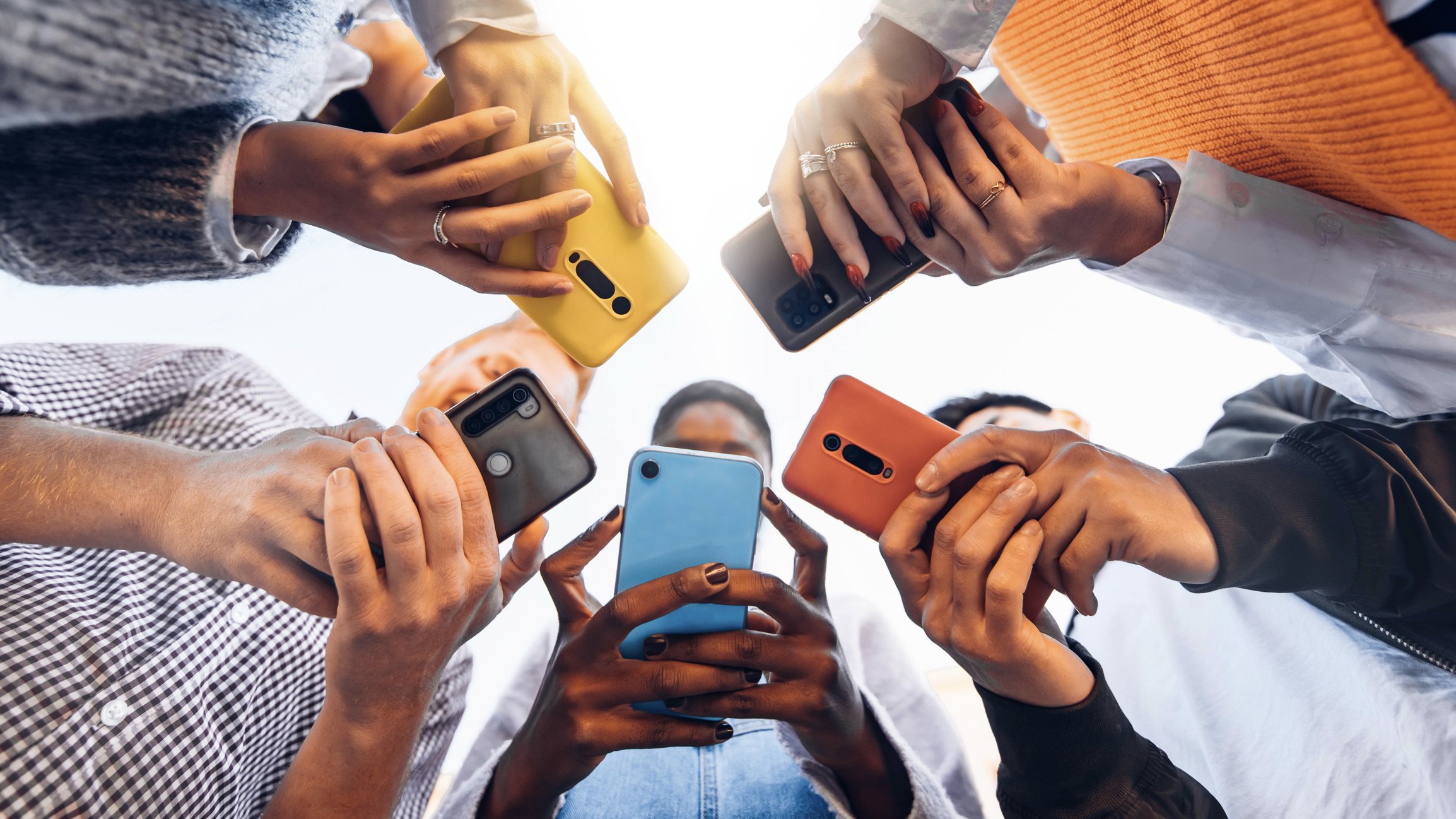 Several people holding smartphones and taking photos or selfies from below, with their hands and phones visible, against a bright sky background.