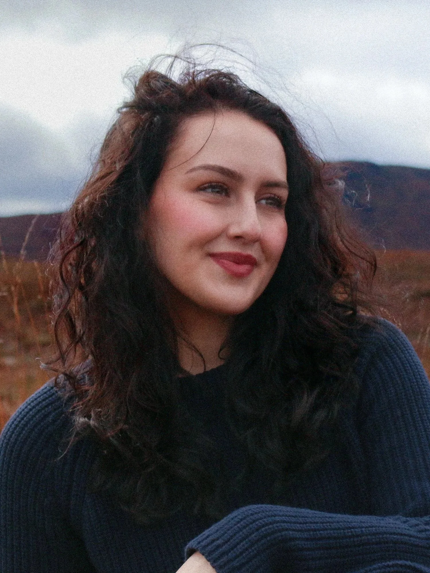 A woman with curly dark hair and light skin smiling outdoors, wearing a navy blue sweater, with a mountain landscape and cloudy sky in the background.