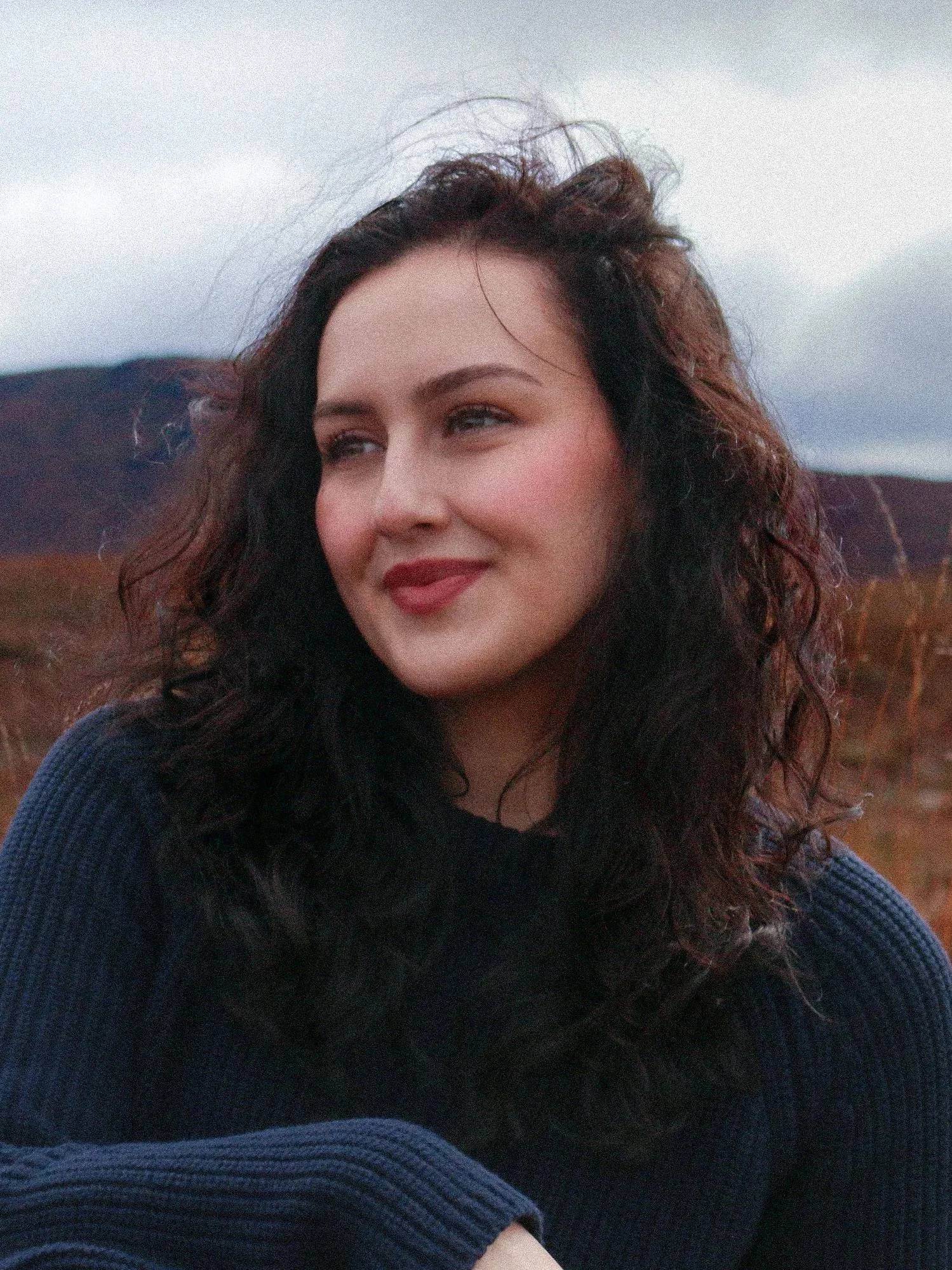 A woman with curly dark hair smiling and looking to her right, wearing a dark blue sweater, outdoors with mountains and cloudy sky in the background.
