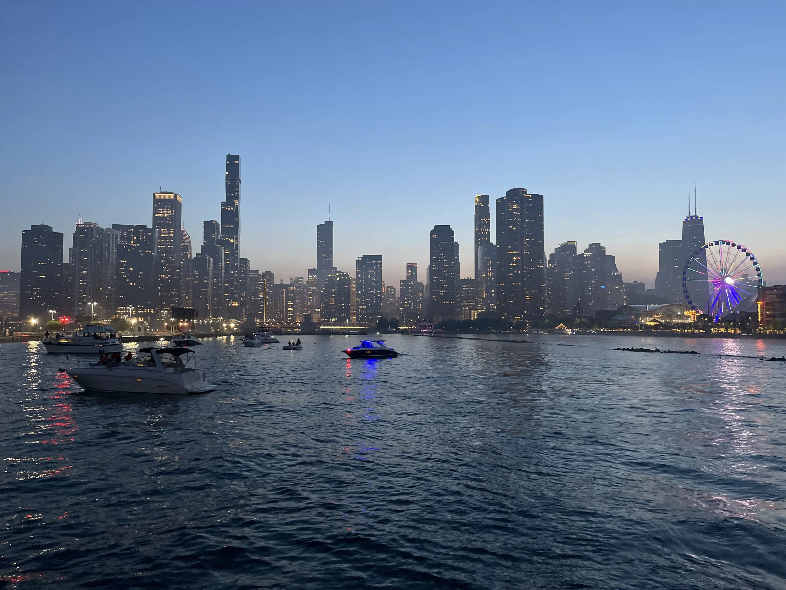 A city skyline at dusk with tall buildings, a Ferris wheel illuminated with purple lights, and boats on the water in the foreground.