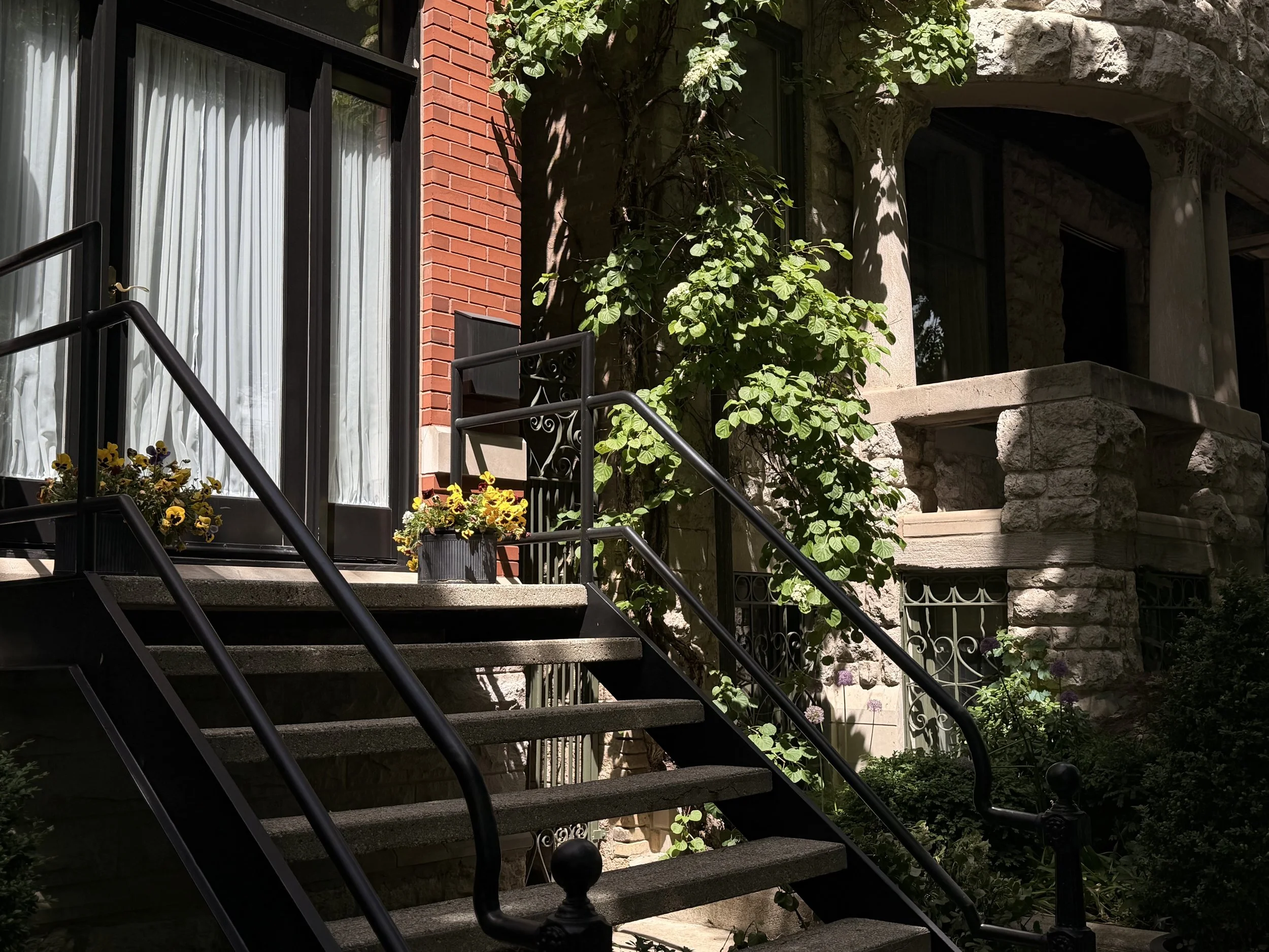 Front porch of a house with brick and stone exterior, black metal staircase, potted yellow flowers, green foliage, and a rounded stone balcony with ornate columns.