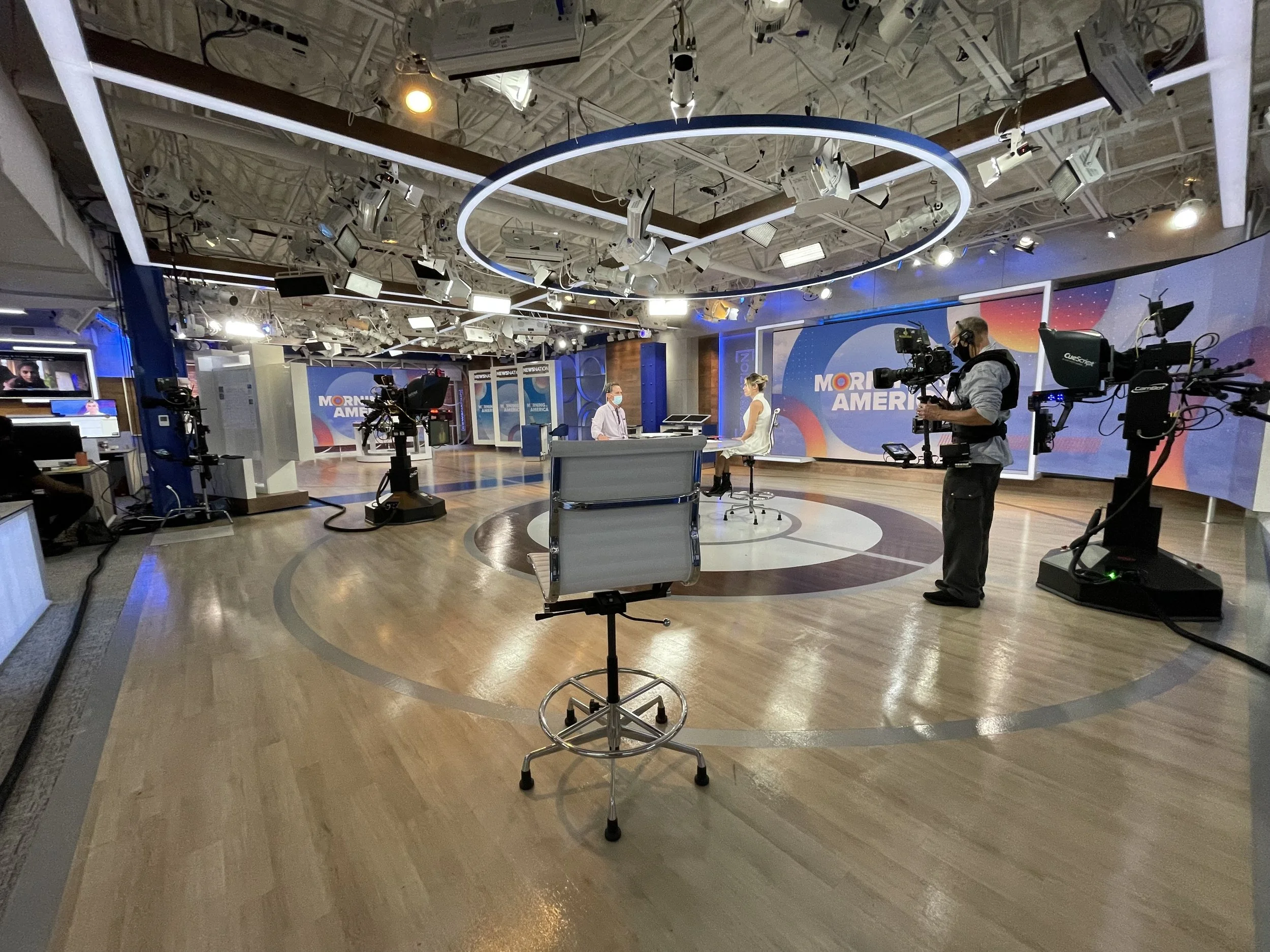 A television studio with lighting equipment and cameras. Two people are seated at a desk, wearing face masks, in front of a backdrop that reads 'Morning America'.